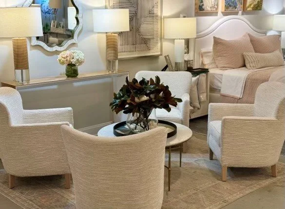 Living room with four beige upholstered chairs surrounding a small round white coffee table, a bed with beige pillows, wall-mounted art, and lamps on a white console table with a flower vase.
