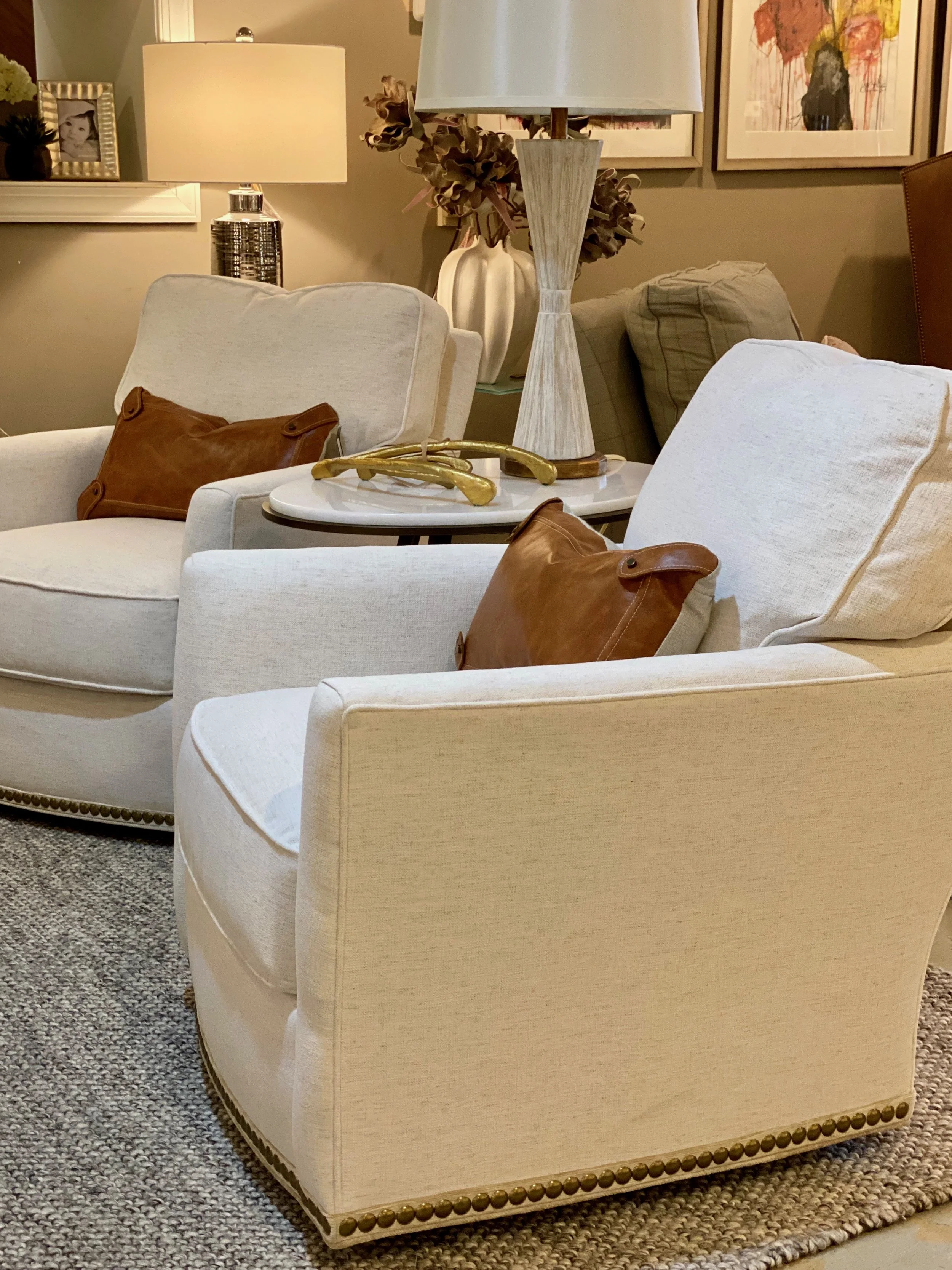 A living room with beige upholstered armchairs, brown pillows, a white side table, a large lamp, and framed artwork on the wall.