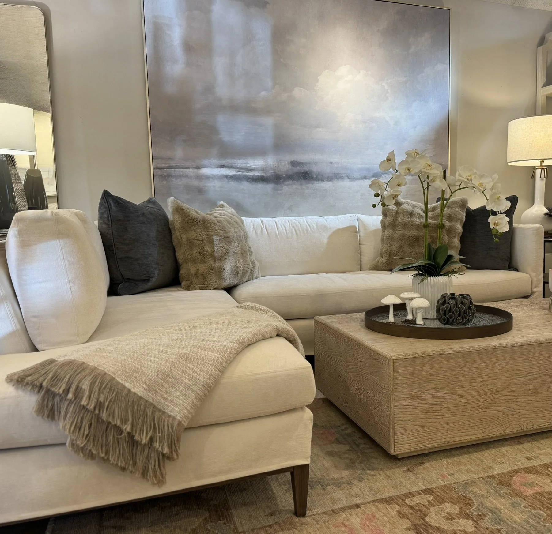 Living room with beige sofa decorated with multiple pillows, including black, beige, and faux fur pillows. A fringed beige throw blanket is draped over the sofa arm. There is a wooden coffee table with a tray holding white decorative mushrooms, black