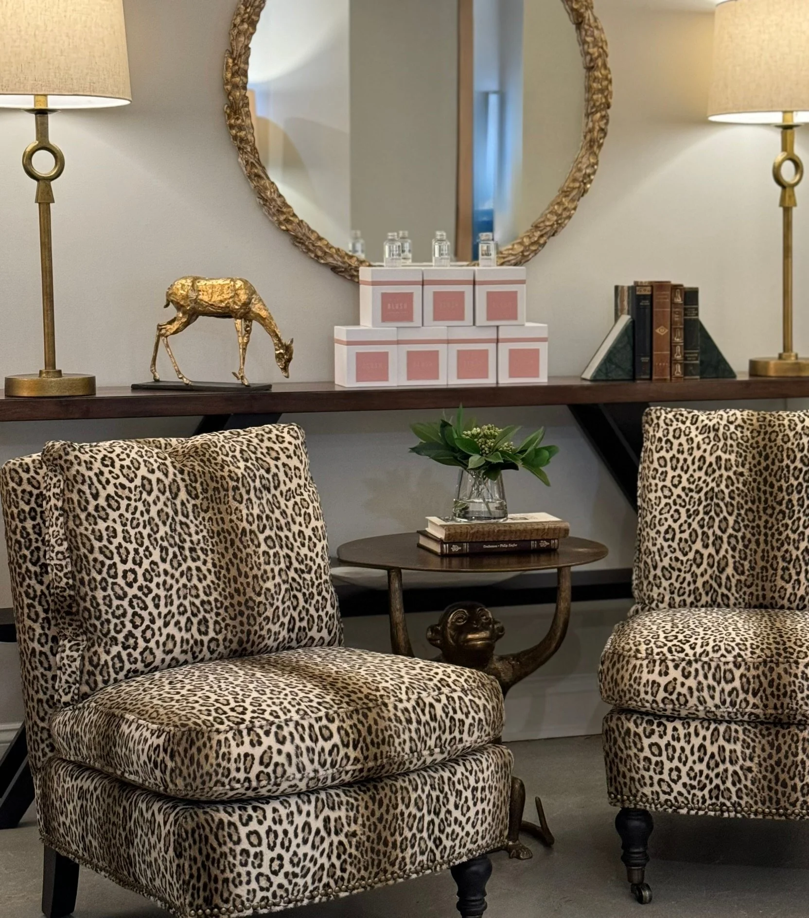 Living room decor with leopard print upholstered chairs, a round wooden table with a glass vase of green plants, and a mirror with a gold textured frame, flanked by two gold lamps with beige shades