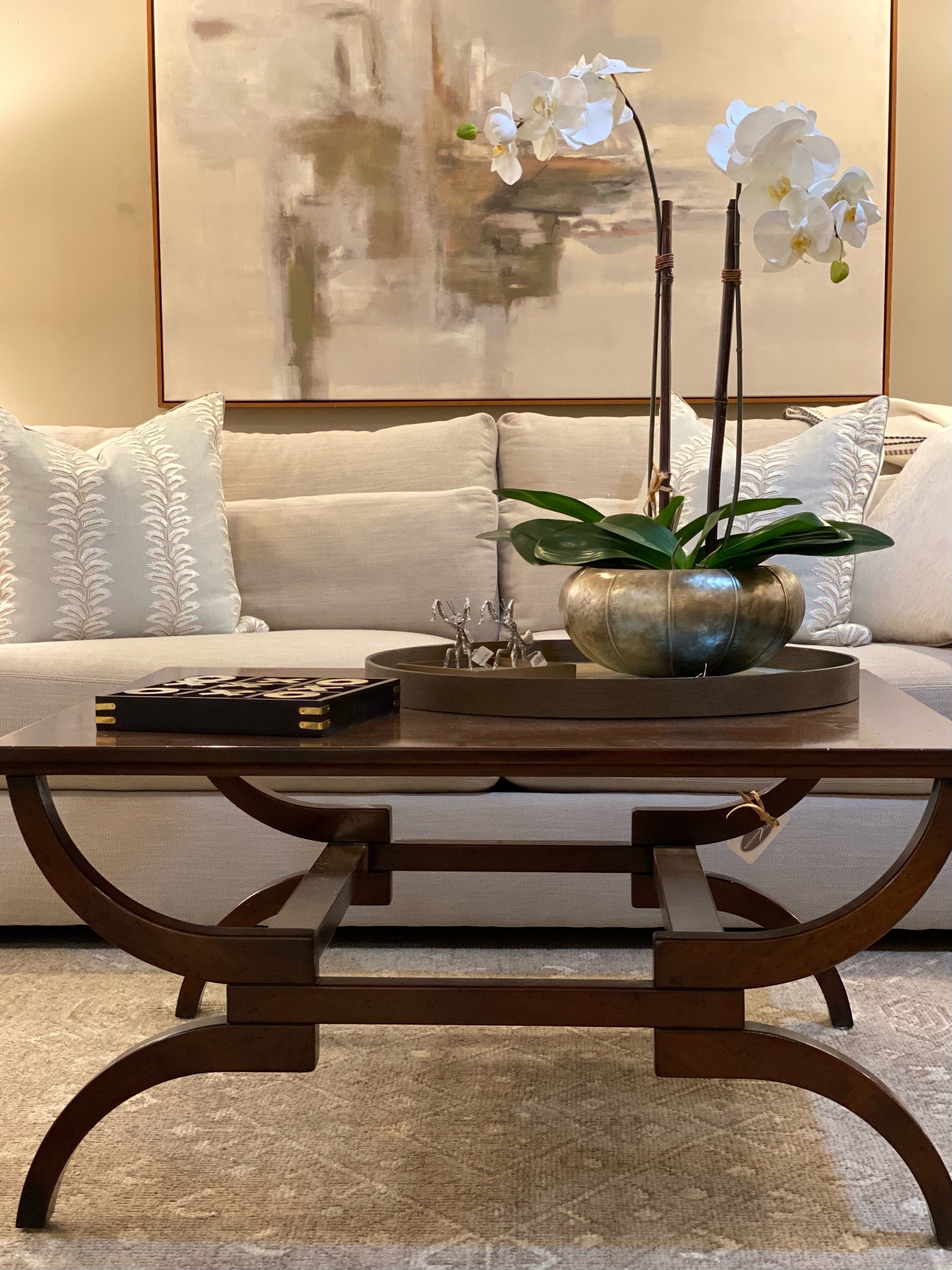 Living room with a beige sofa decorated with white embroidered pillows, a wooden coffee table with a decorative tray holding small metal animal figurines, a large gold-colored vase with white orchids, a round tray beneath the vase, an abstract landsc