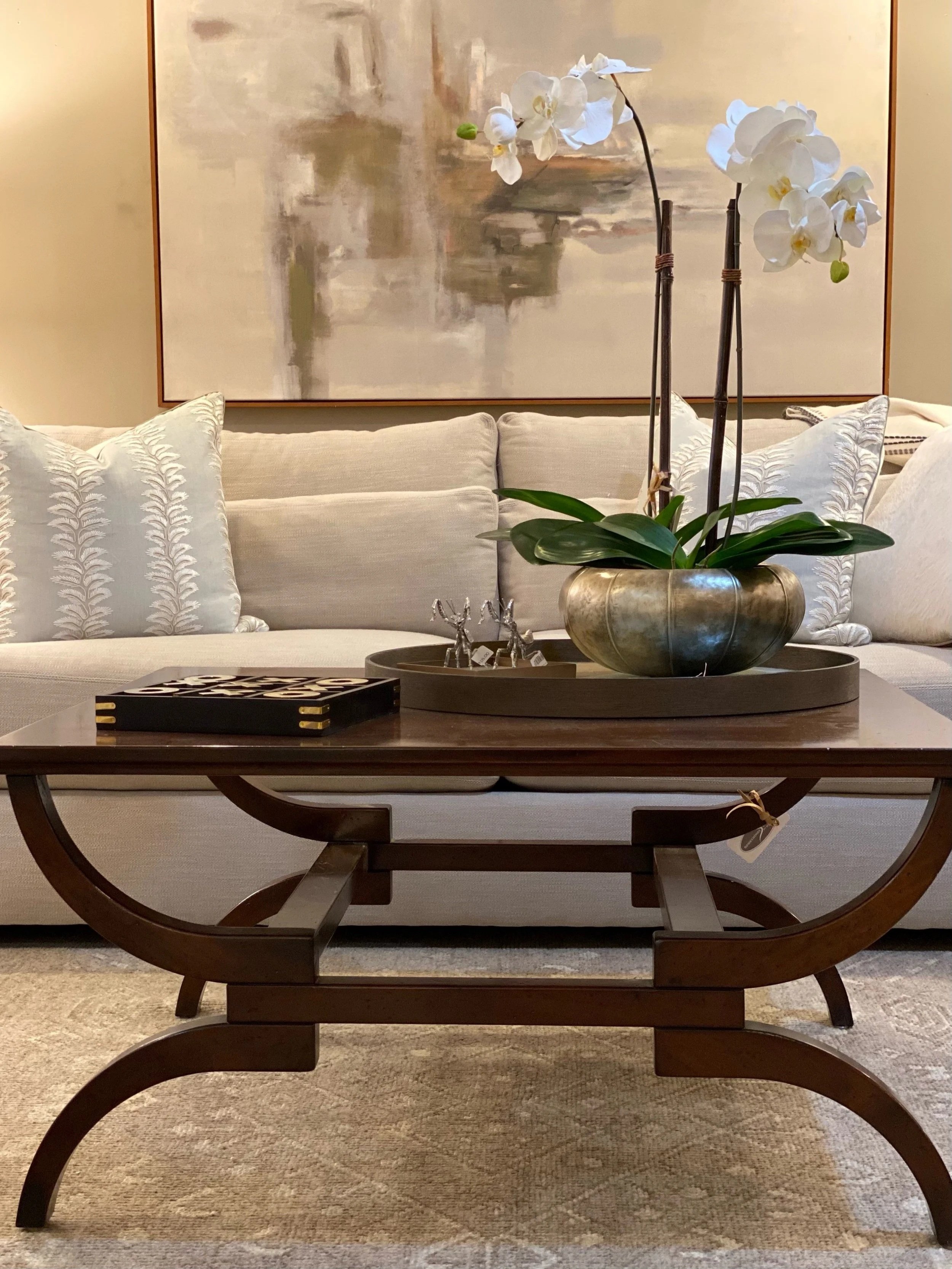 A living room with a beige sofa and patterned pillows, a wooden coffee table with a tray holding small reindeer figurines, a decorative orchid plant in a brass pot on the table, a large abstract painting on the wall behind the sofa, and a beige rug on the floor.