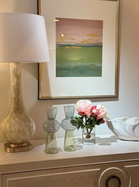 Decorative bedroom dresser with a large beige table lamp, two small glass vases, a small glass jar with pink peonies, and a white ceramic bowl, with a framed landscape painting hanging above.
