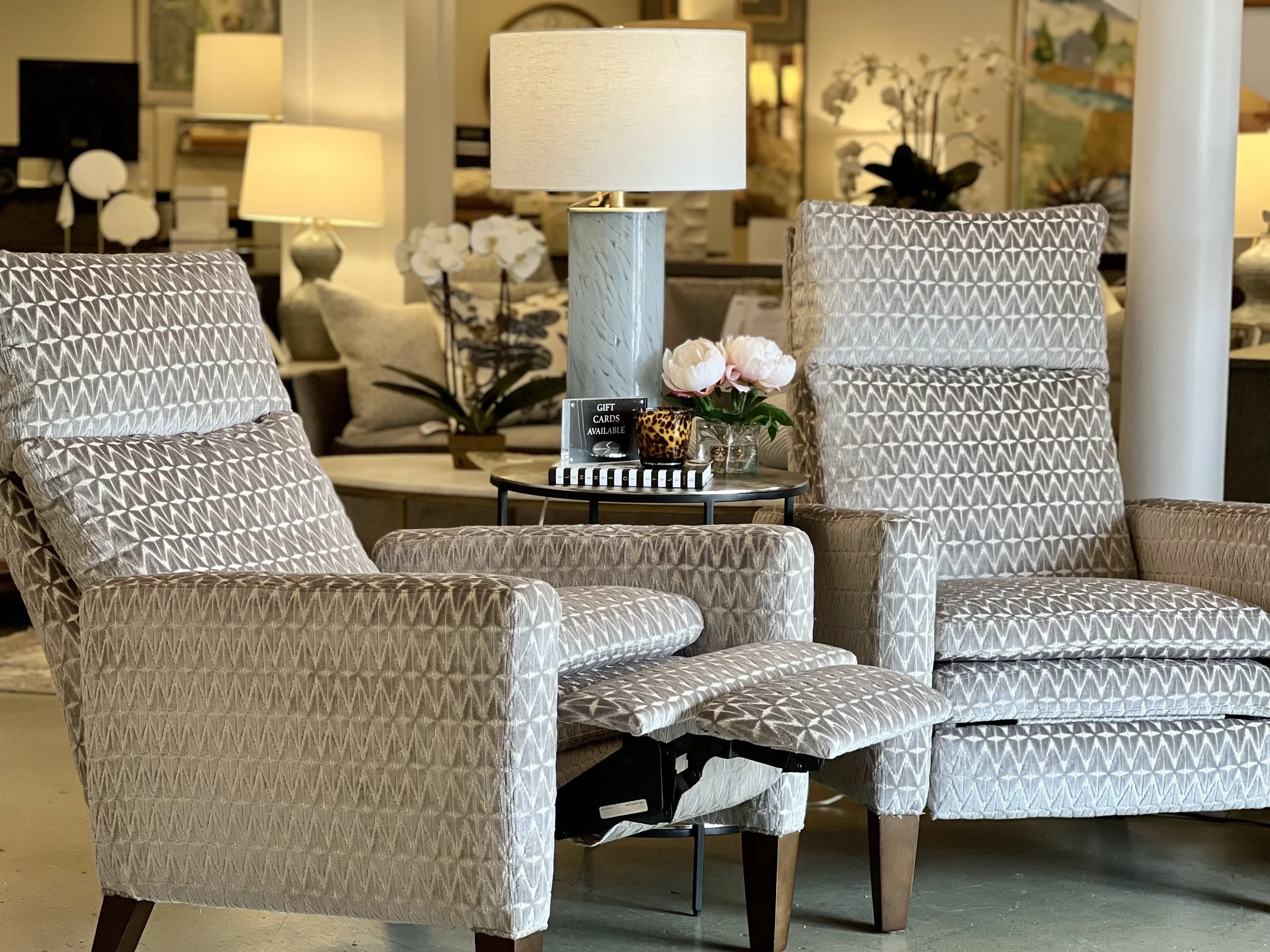 Showroom with patterned armchairs, side table with pink flowers, lamps, and various home decor items in the background.