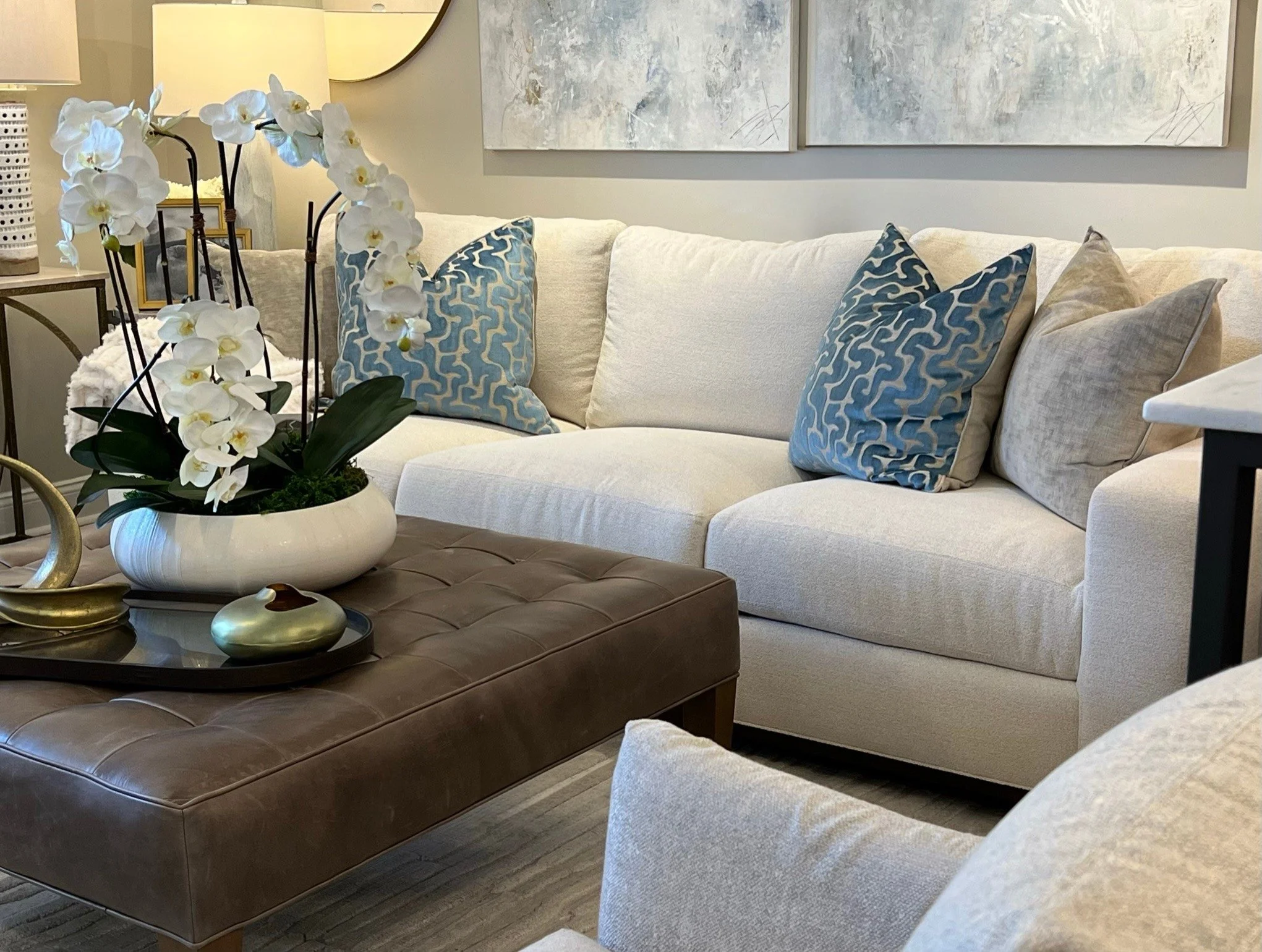 Living room with a beige sofa, blue patterned pillows, a plush beige chair, a dark brown tufted coffee table, and a decorative floral arrangement of white orchids in a white bowl on the coffee table.