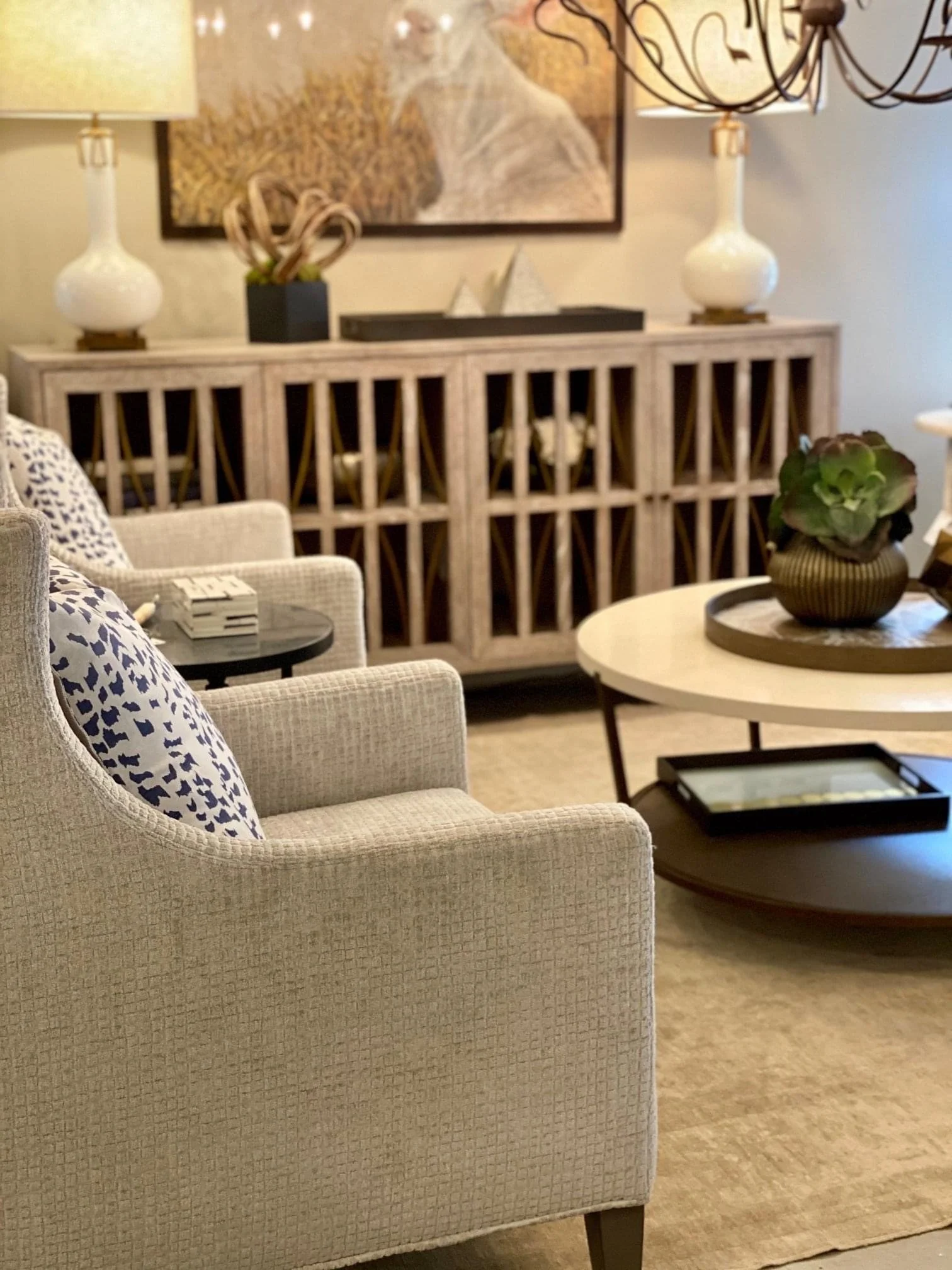 Living room with beige armchair with blue and white patterned cushion, round black side table with white box, modern beige textured sideboard with decor items, black tray with a plant, white round coffee table with a black tray, and beige carpet. The
