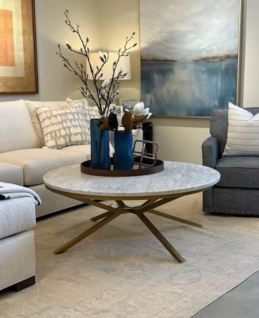 Living room with a beige sofa, a gray armchair, and a round marble coffee table with a wooden base. On the table is a tray with two blue vases and a decorative branch with white flowers. A large abstract painting hangs on the wall behind the sofa.