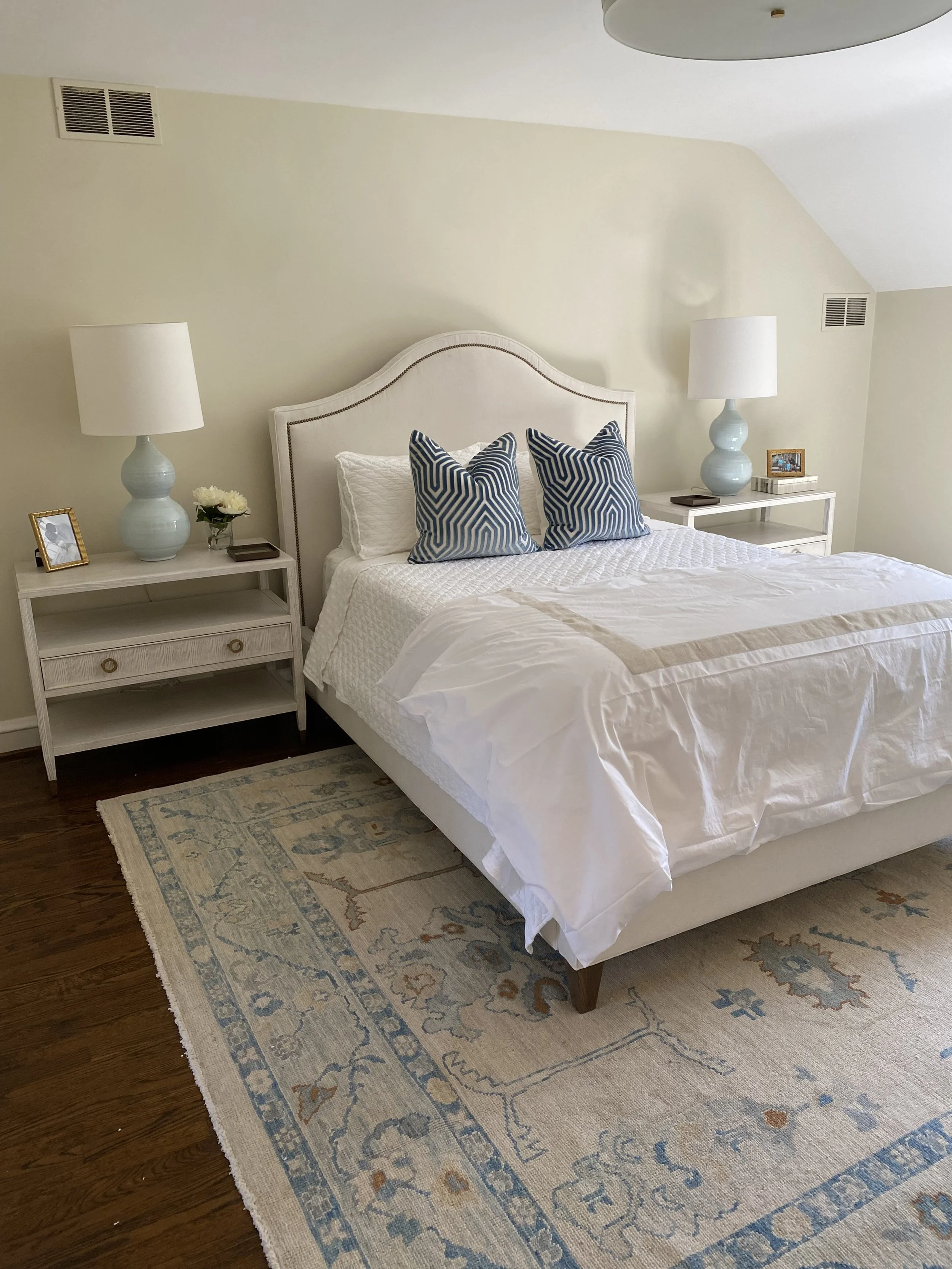 A bedroom with a white upholstered bed, two nightstands with white lamps, and blue patterned pillows. There is a framed photo, a flower vase, and a book on each nightstand. The room has beige walls, a patterned area rug, and wooden floor.