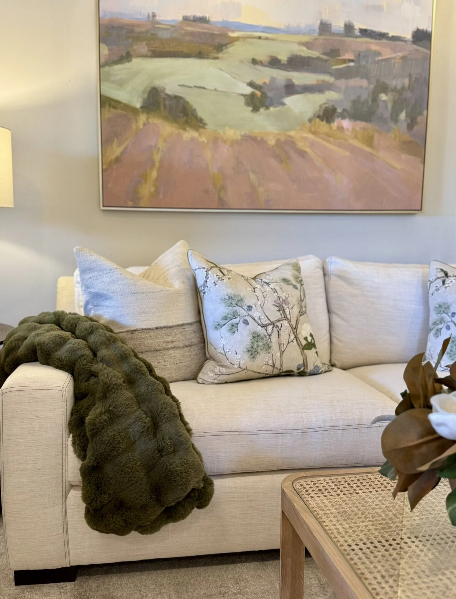 A beige sofa decorated with decorative pillows, a textured fur throw blanket, and a green pillow with an embroidered floral design. A large framed landscape painting hangs above the sofa. To the right, a wooden side table with a rattan top holds a bo