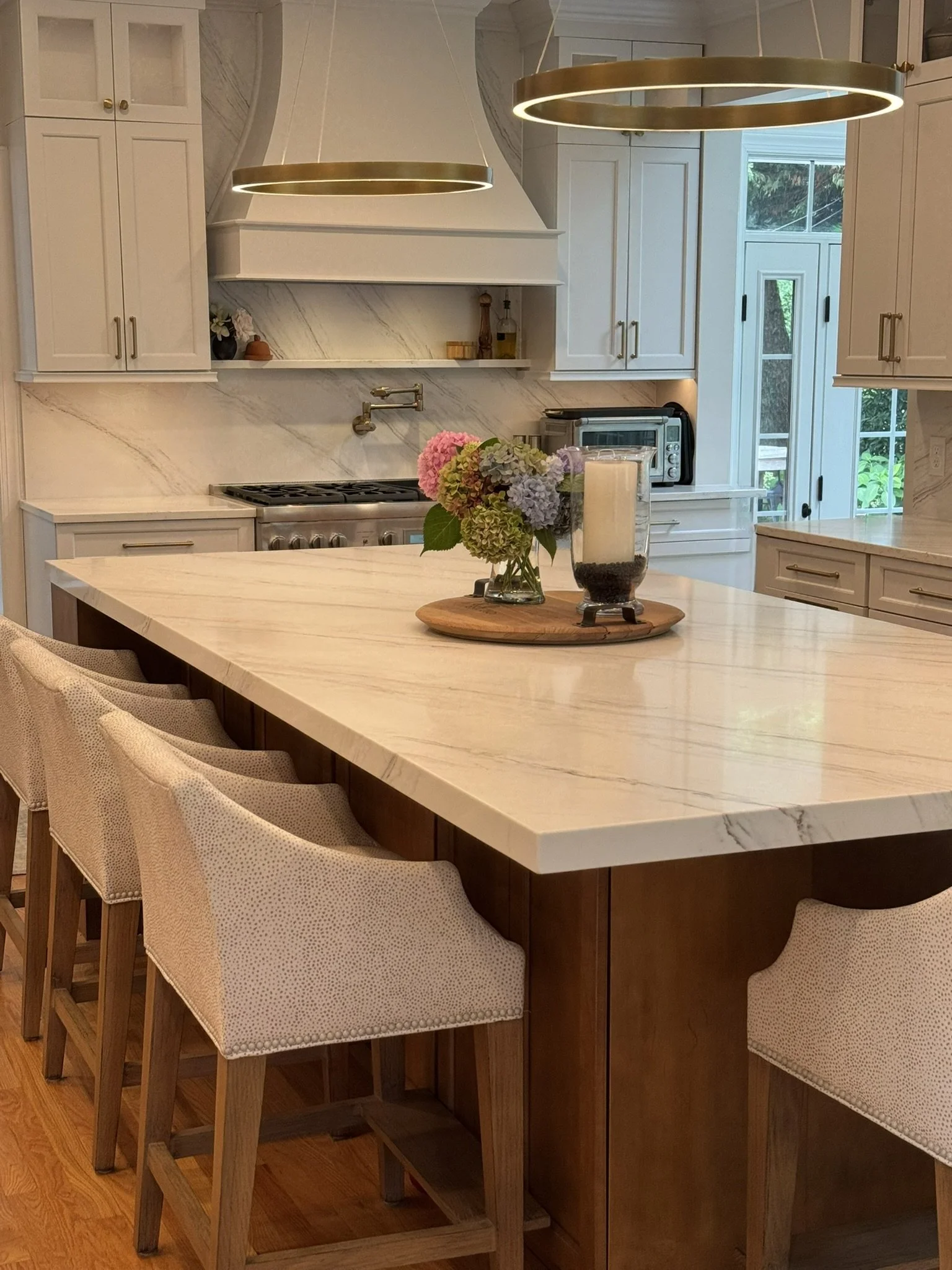 A kitchen with white cabinets, a marble backsplash, and a large island with a wooden base. Four upholstered bar stools are lined up at the island. There is a flower arrangement and a candle on a wooden tray on the counter. Modern pendant lighting hangs above the island. In the background, there are windows and a door leading outside.