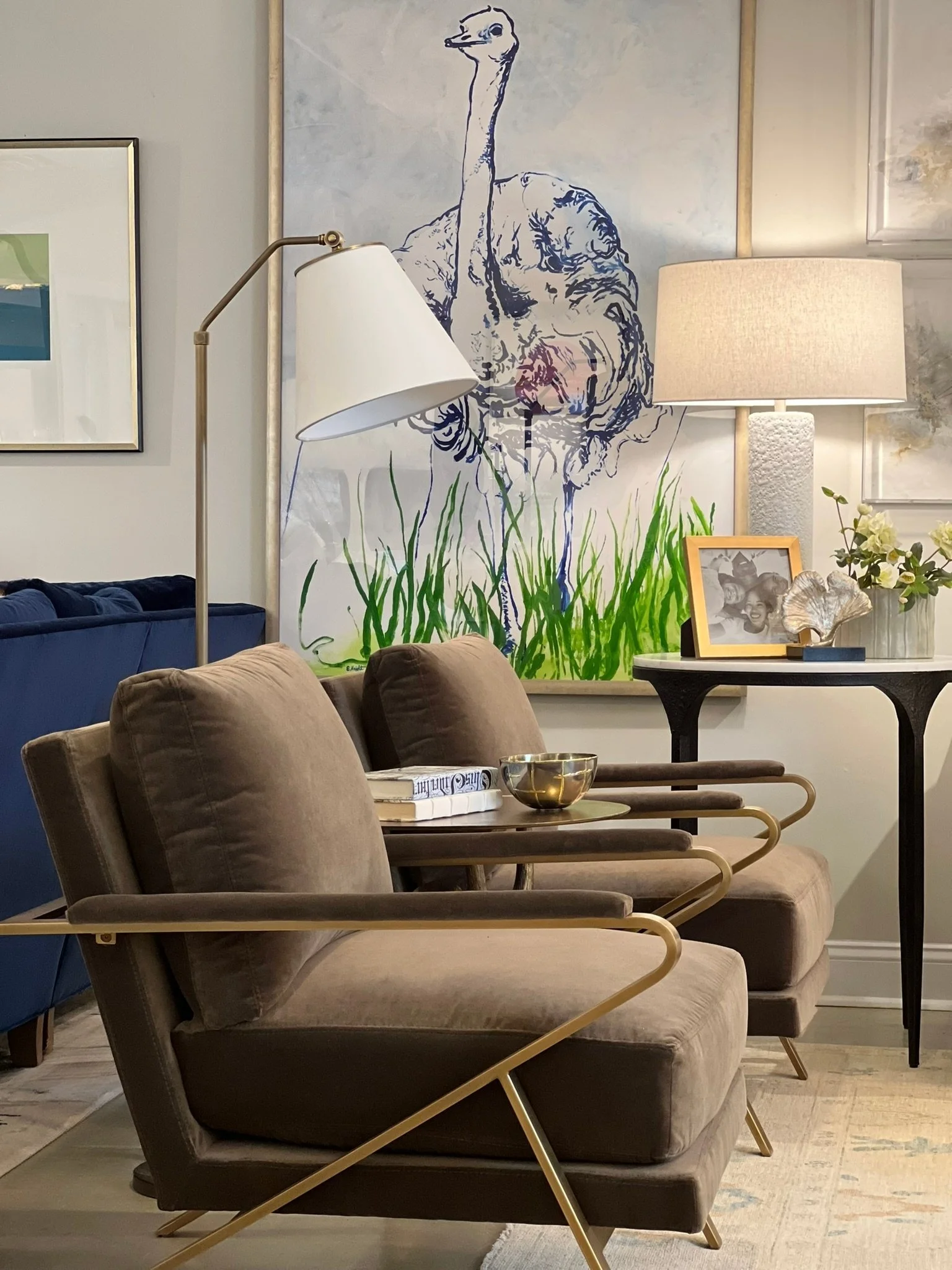 A cozy living room with brown velvet armchairs, a round table with a framed photo and decorative items, a large lamp, and a wall art of an ostrich with grass at the bottom.