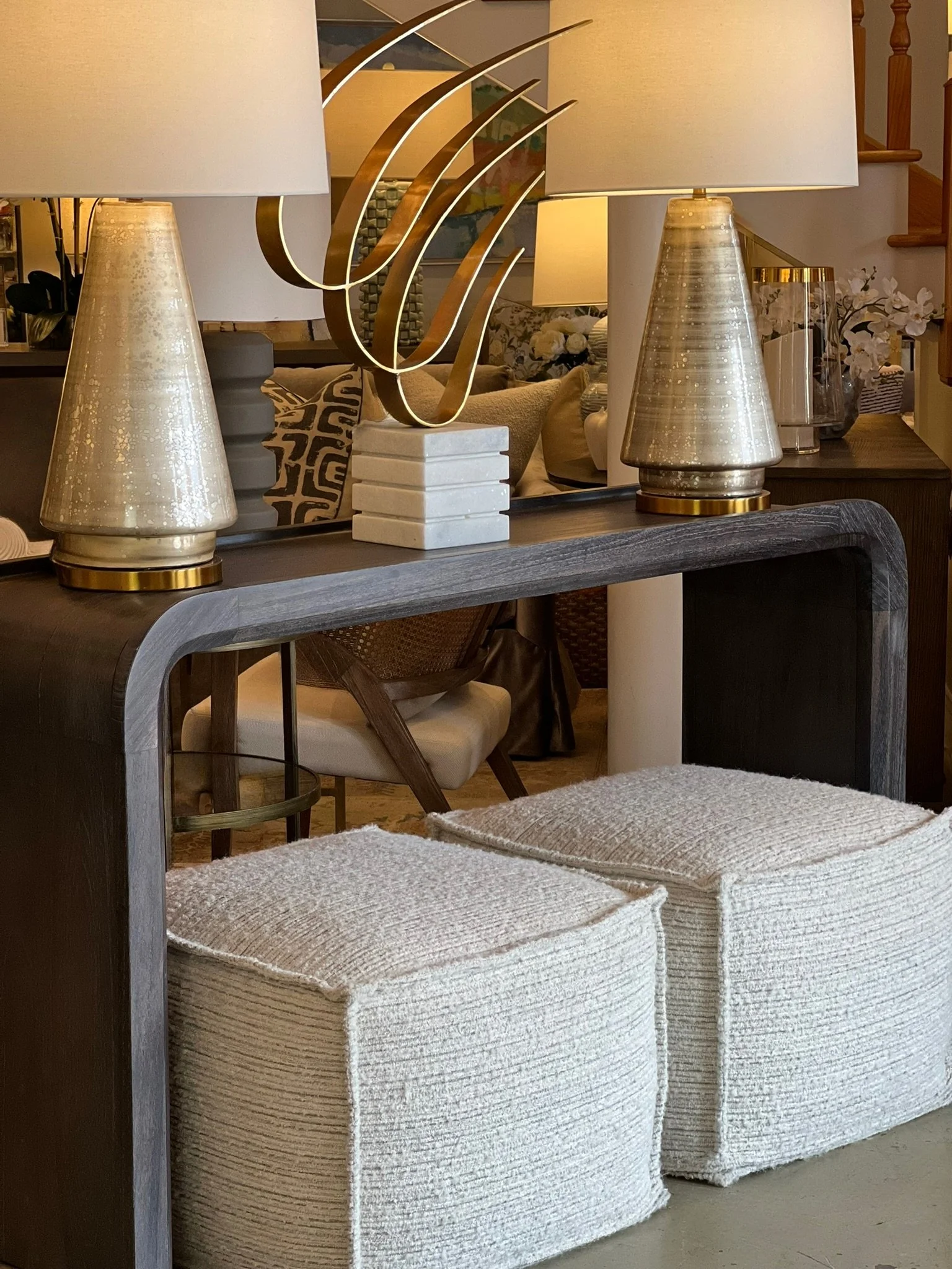A dining room with a dark wood table, cream-colored upholstered chairs, two table lamps with shiny metallic bases, a mirror reflecting a decorative sculpture, and various decorative items including flowers and pillows.