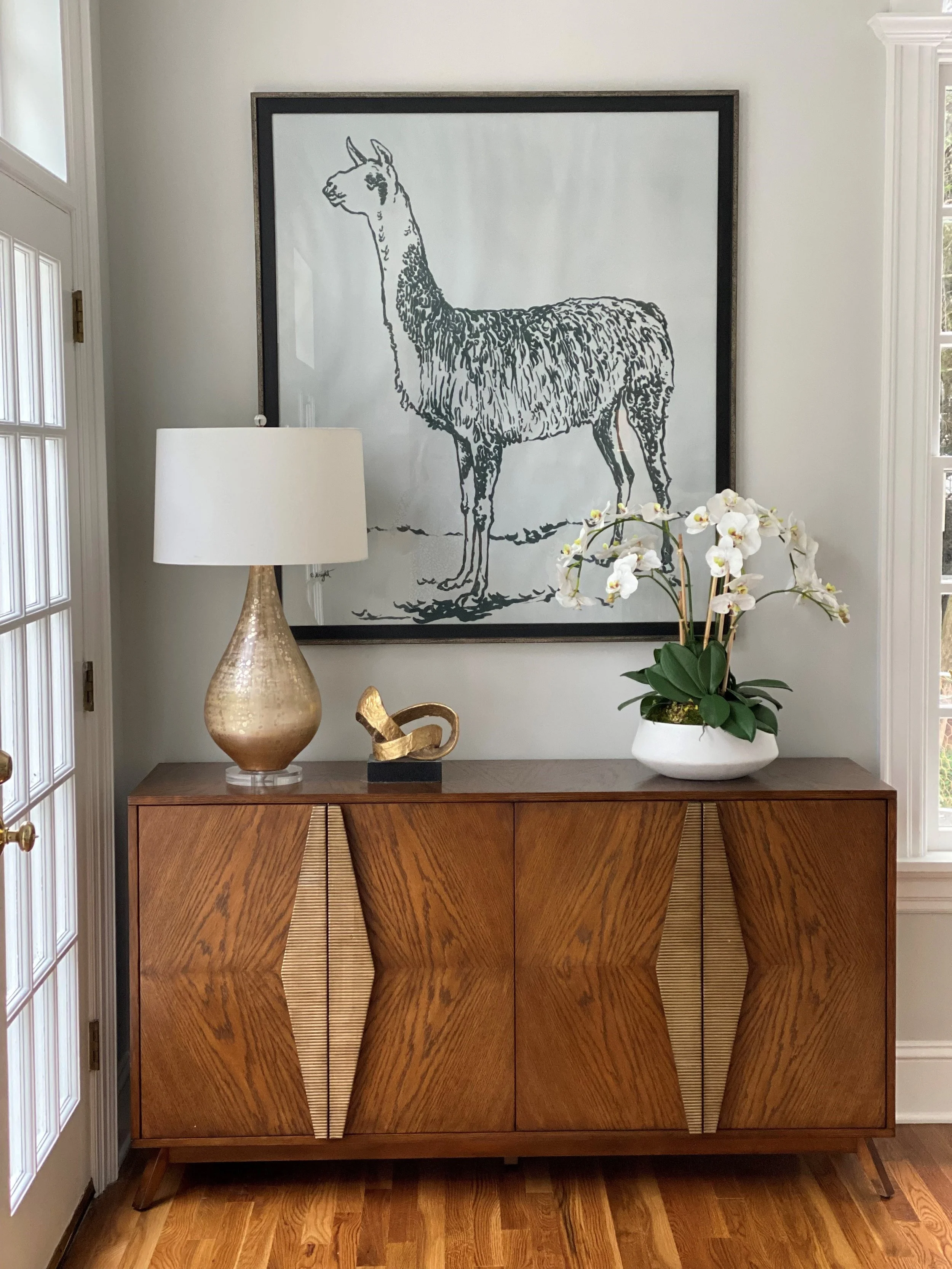 A wooden sideboard with a geometric pattern on its doors, a white table lamp with a gold base, an abstract gold sculpture, a potted white orchid plant, and a large framed black-and-white drawing of an llama on the wall behind.