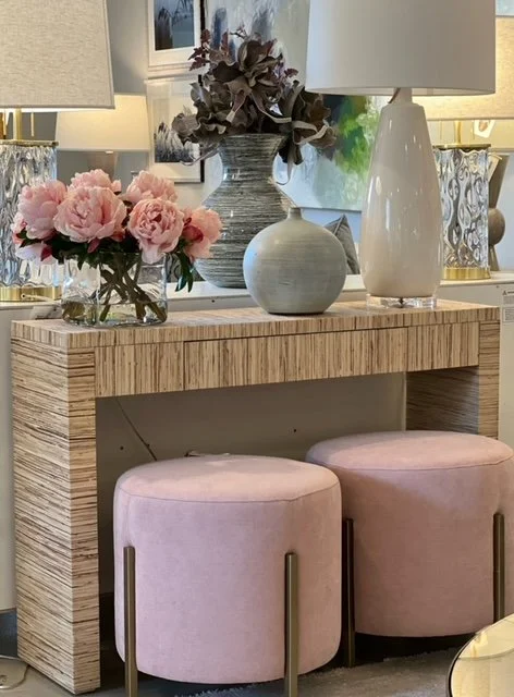 A wooden console table with decorative vases and lamps on it, flanked by two pink velvet ottomans with gold legs.