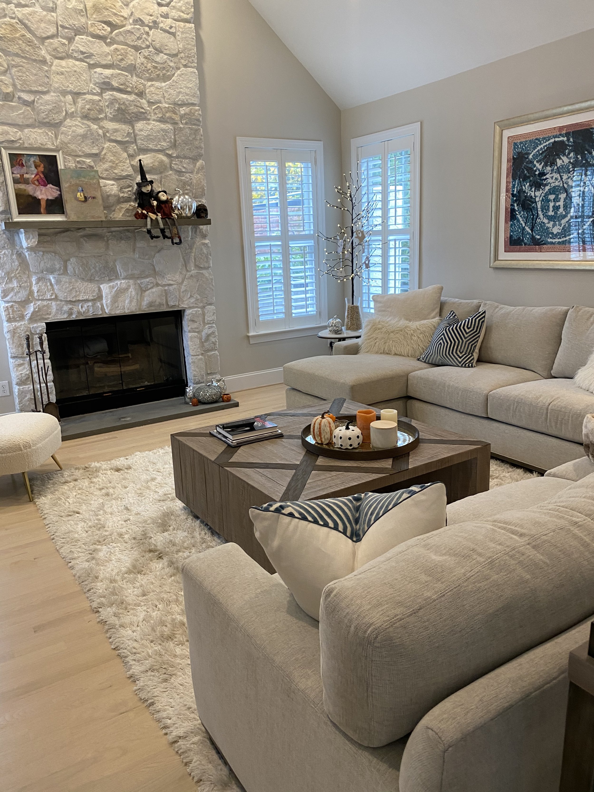 Living room with beige sectional sofa, white shag rug, wooden coffee table with fall-themed decorations, stone fireplace, picture frames, holiday dolls on mantel, floor lamp with branches and LED lights, two windows with white shutters, framed artwork on wall.