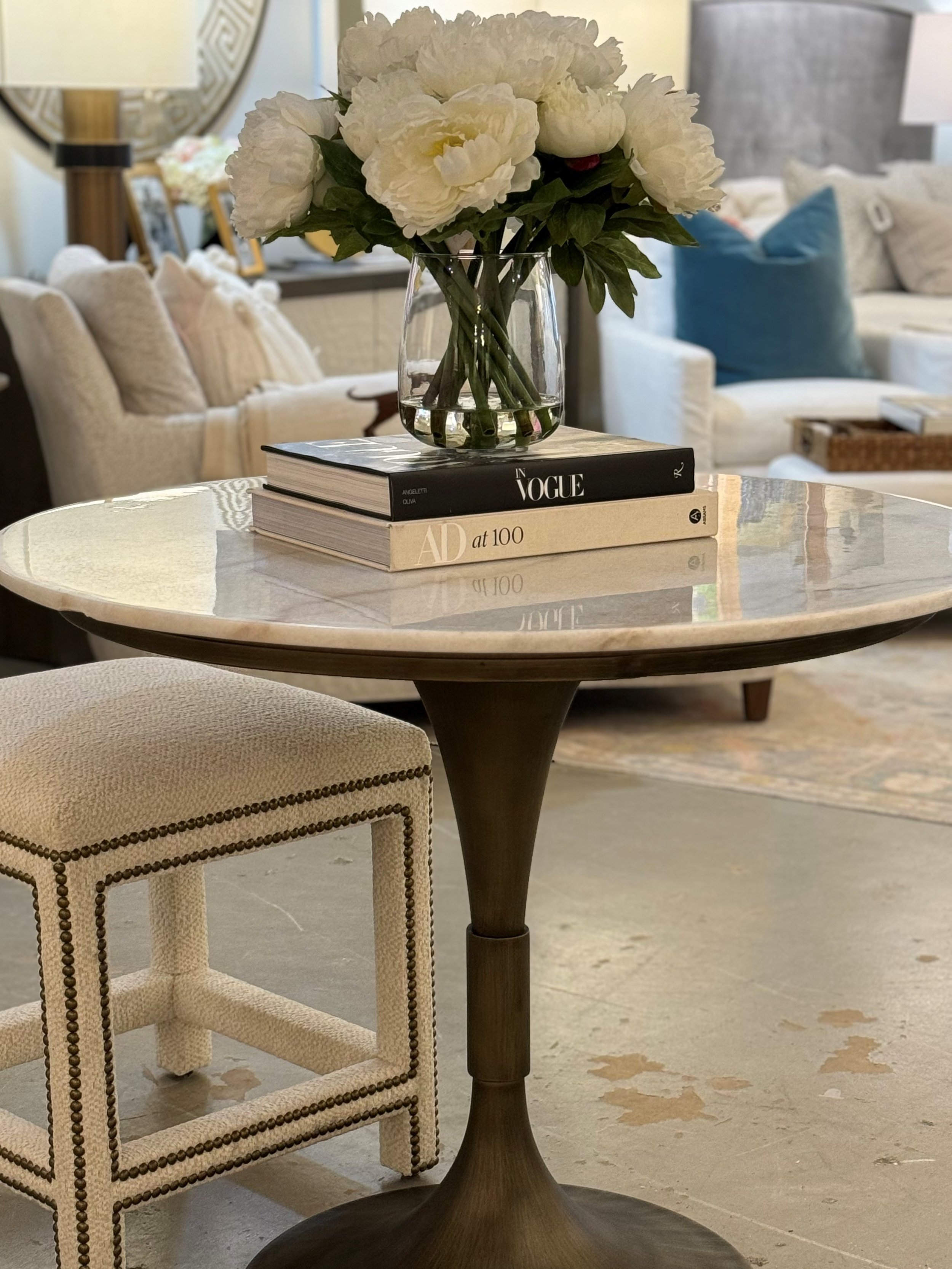 A clear glass vase with white peony flowers and green leaves, placed on top of two books on a round wooden table in a living room.
