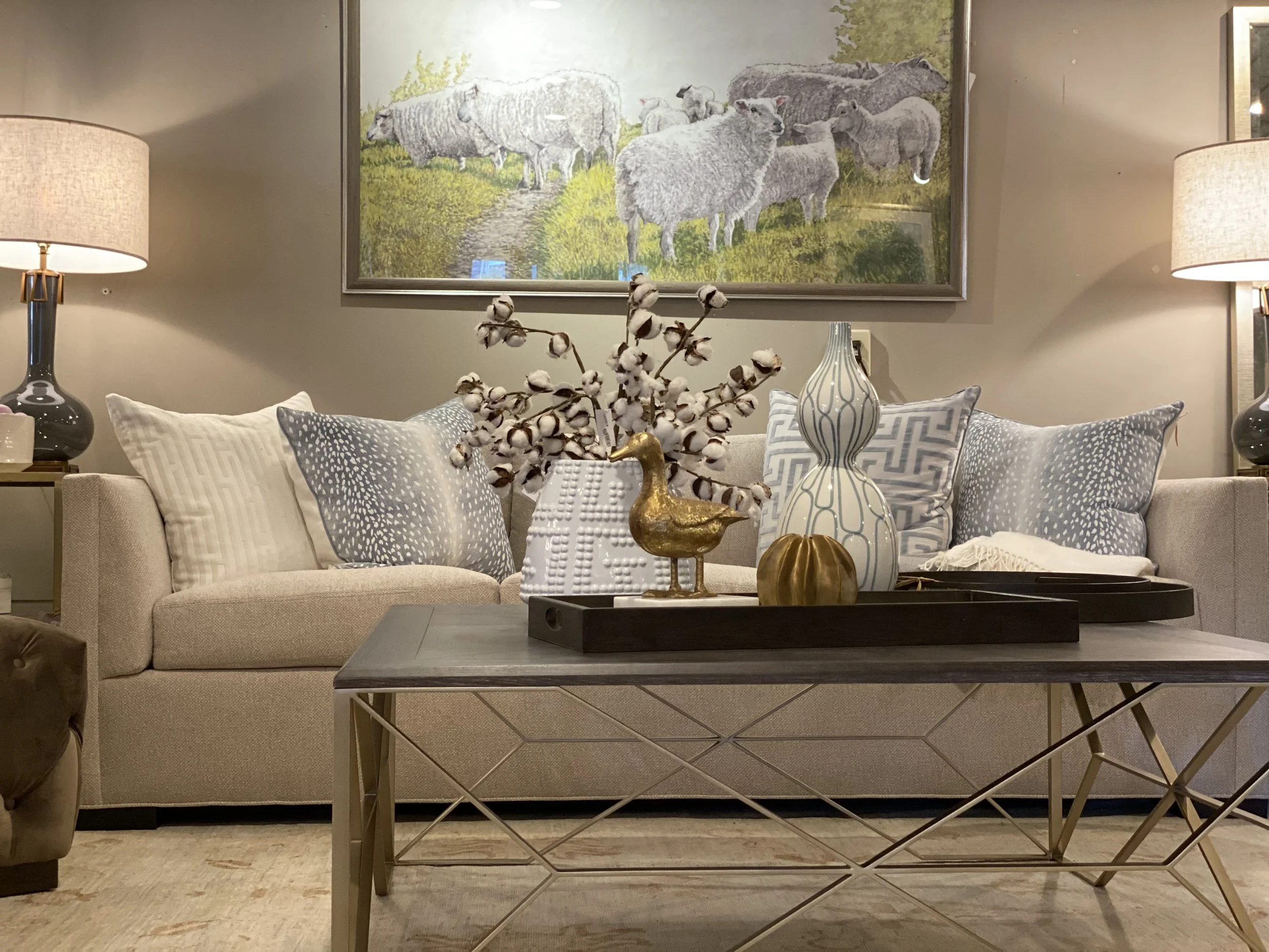 Living room with beige sofa, decorative pillows, coffee table with cotton branches and vases, framed sheep picture on wall, two table lamps.