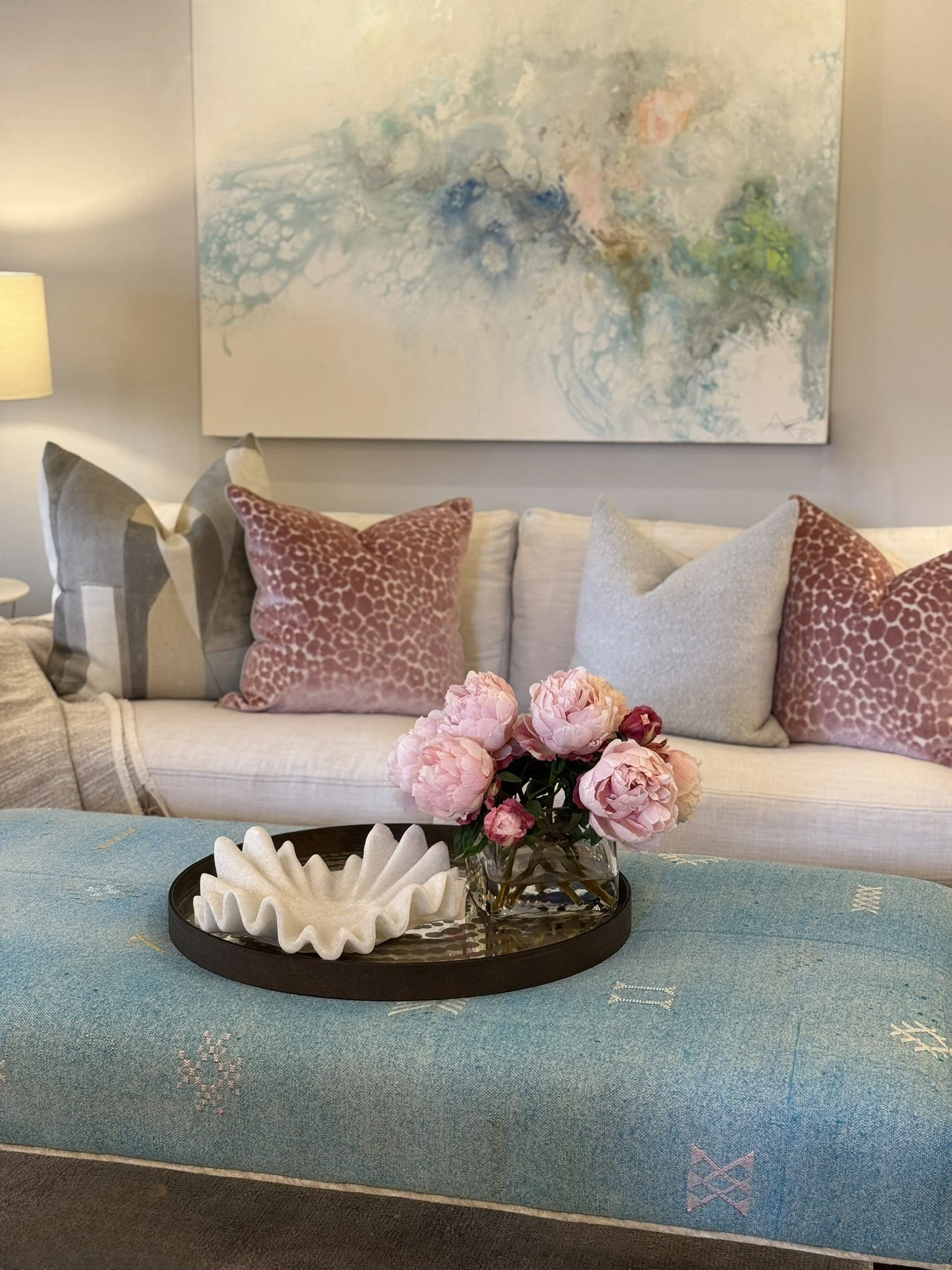 Living room with a beige sofa, decorative pillows, a glass vase with pink flowers, a black tray, and an abstract painting on the wall.