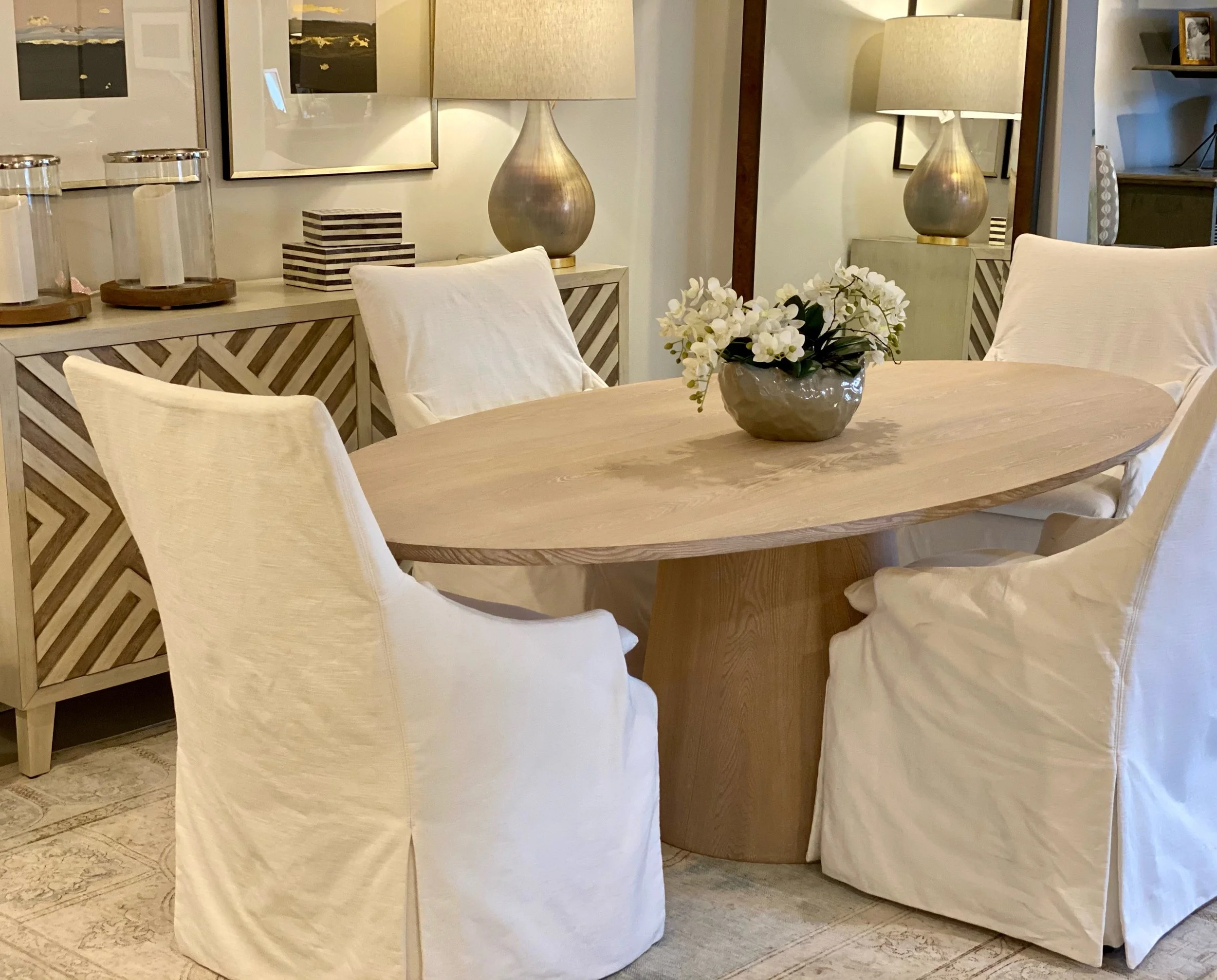 Interior of a dining room with a wooden table, white upholstered chairs, a vase with white flowers, two large lamps with beige shades, and framed artwork on the wall.