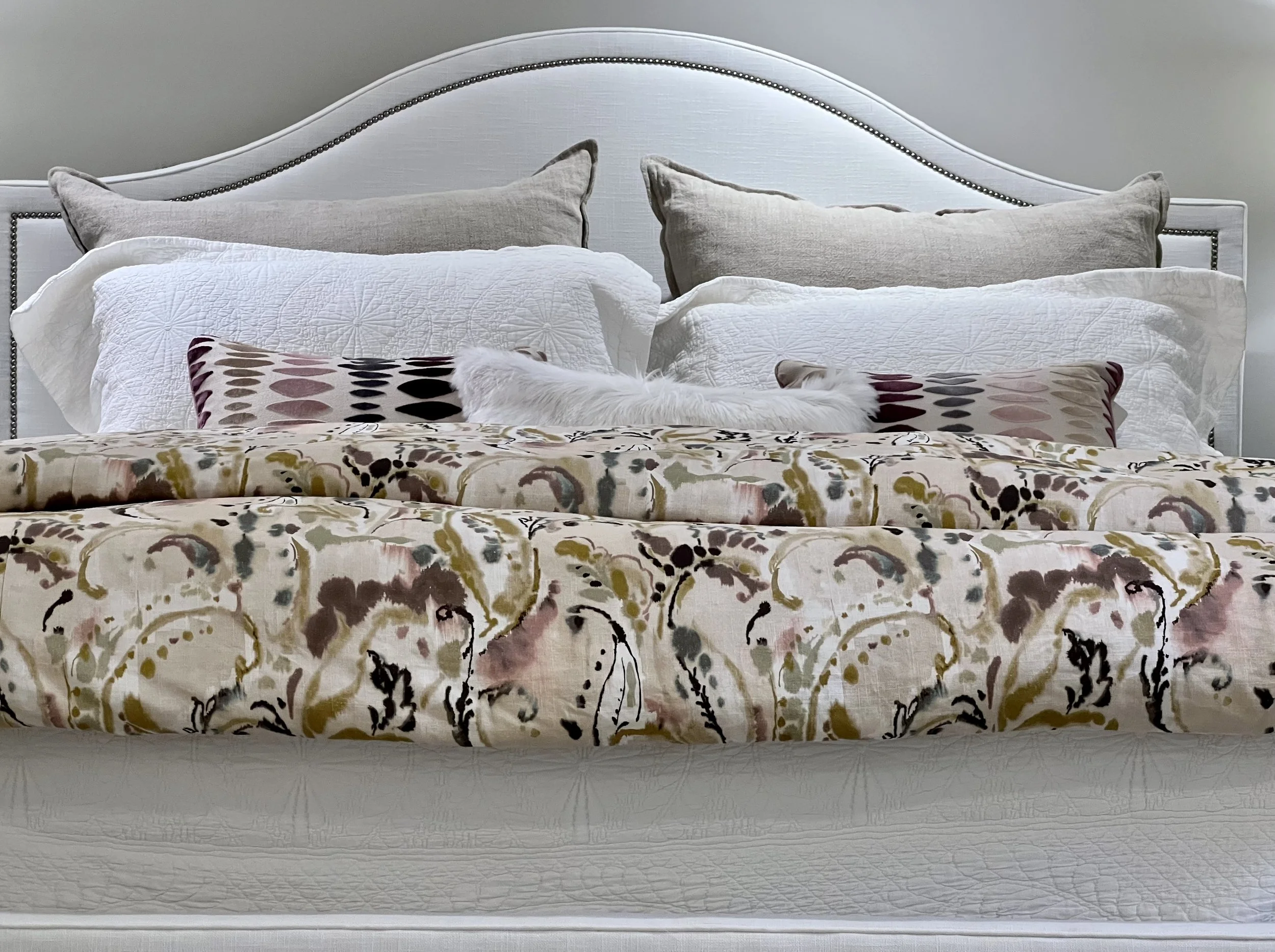 A bed with a textured beige headboard, multiple pillows including beige and white ones, a patterned throw blanket, and a floral bedspread.