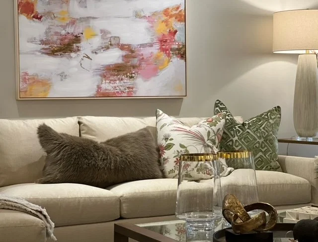 A cozy living room with a beige sofa decorated with a furry brown pillow and three patterned throw pillows. There's an abstract art painting with pink, yellow, and brown tones hanging on the wall above the sofa. A tall table lamp with a beige shade s