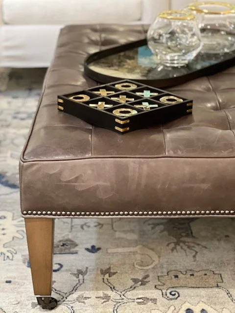 A brown leather ottoman with tufted upholstery and nailhead trim, placed on a patterned area rug.