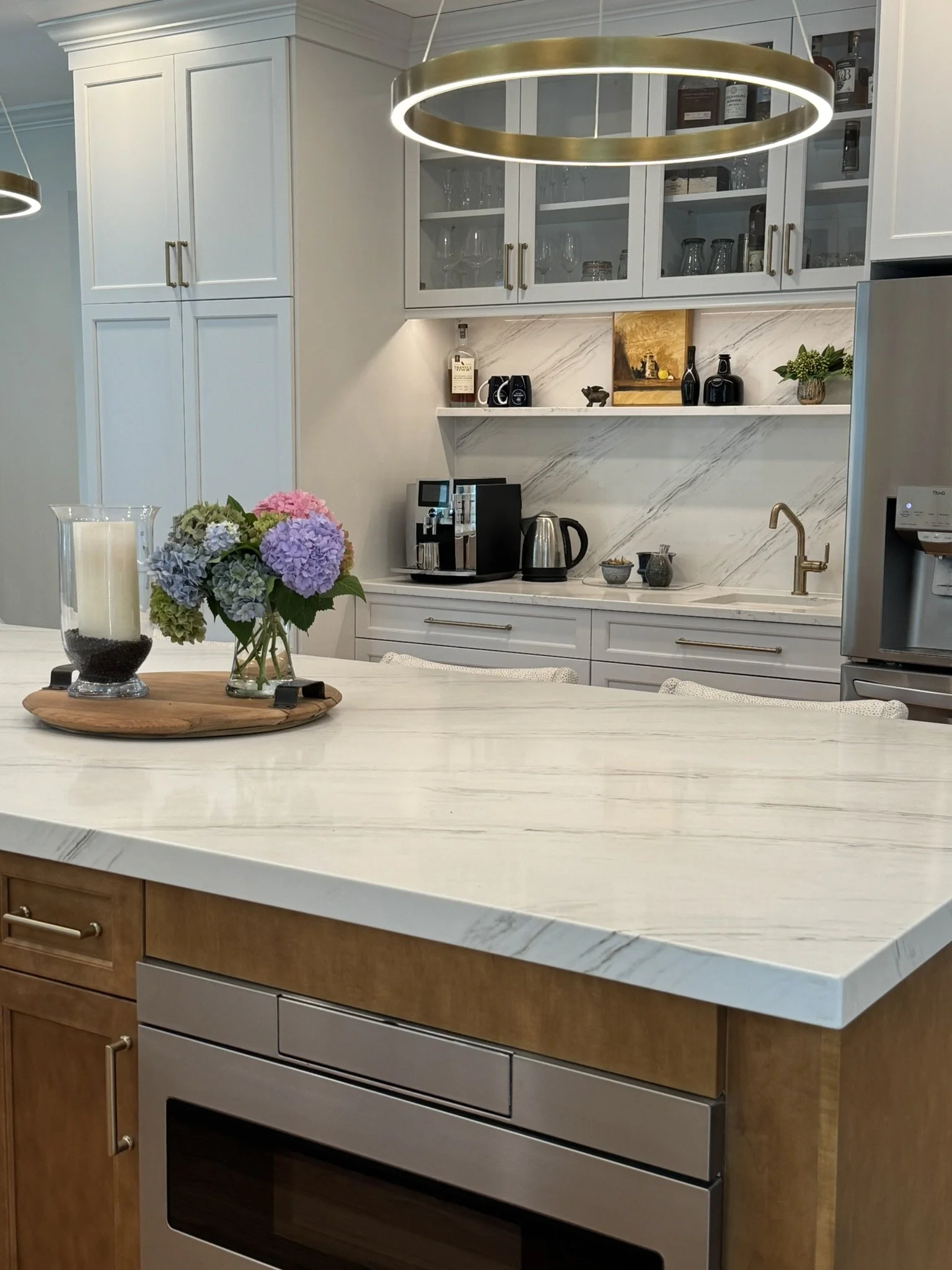 Modern kitchen with white marble countertop and backsplash, light brown and white cabinets, and a built-in oven. There is a small bouquet of hydrangeas on the counter, and various kitchen appliances and decorative items are arranged on shelves.