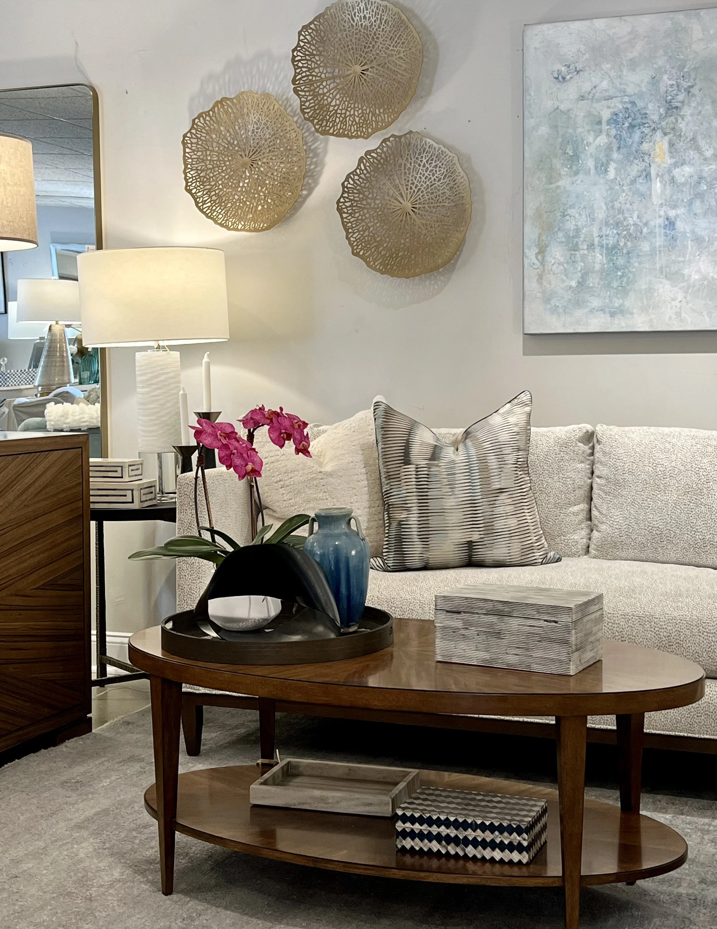 Living room with a white sofa adorned with a striped decorative pillow, standing against a light-colored wall decorated with three gold-tone wall art pieces resembling seashells, a framed abstract painting, two white table lamps with beige shades, a 