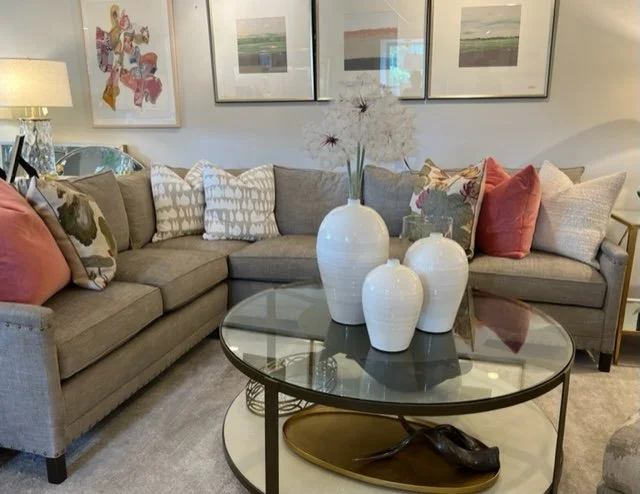 Living room with a beige sofa, decorative pillows, three white vases with floral arrangement on a glass coffee table, colorful framed artwork on the wall, and soft lighting.