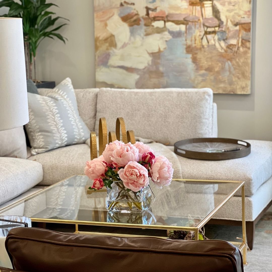 A living room with a beige sofa, a glass coffee table with gold frames, a bouquet of pink peonies in a glass vase, a tray on the sofa, and a large abstract painting on the wall.