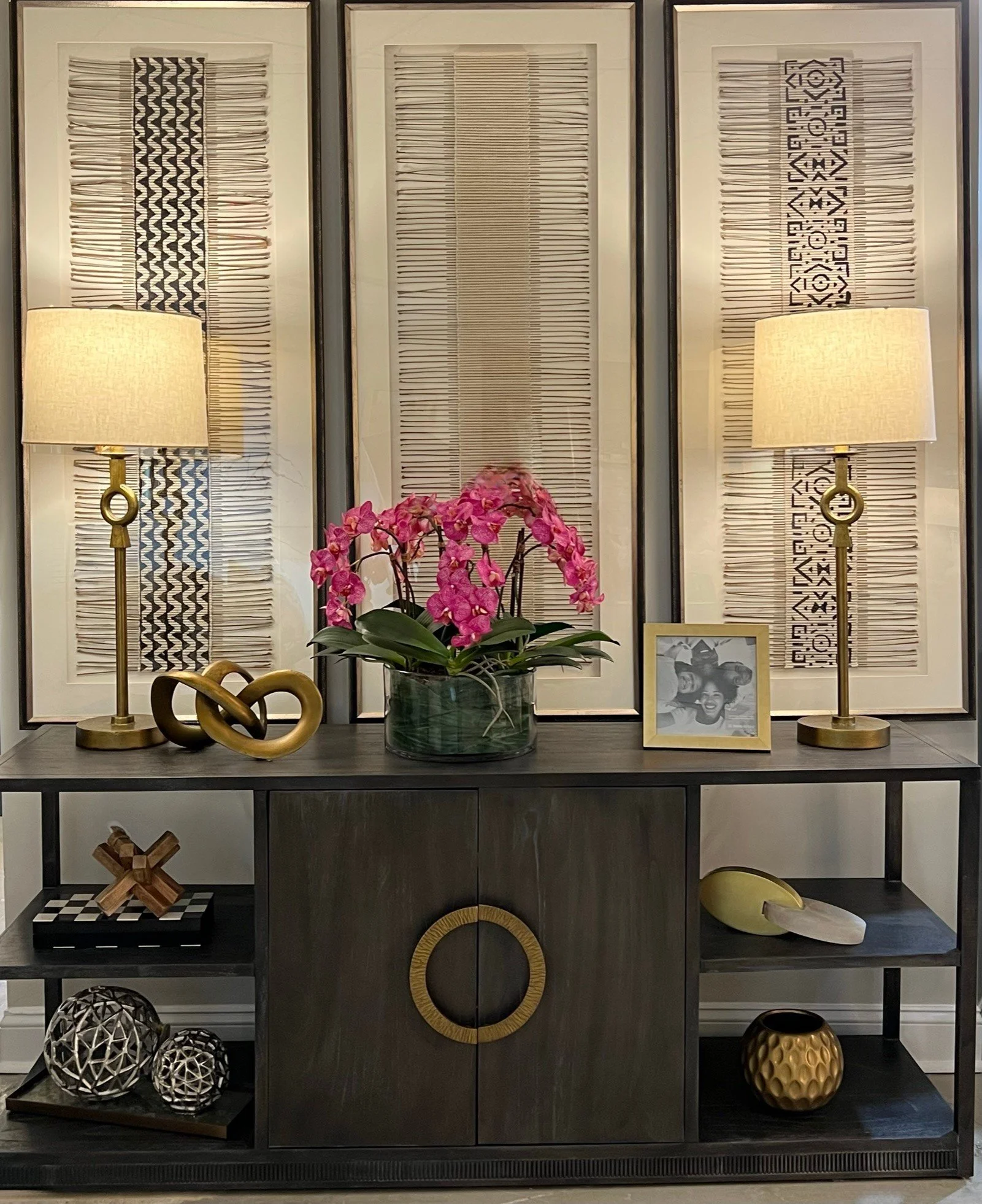 Decorative black and gold display shelf with various geometric and floral ornaments, a vase with pink orchids, a framed black and white photo, two table lamps with white shades, and woven and wooden decorative objects.