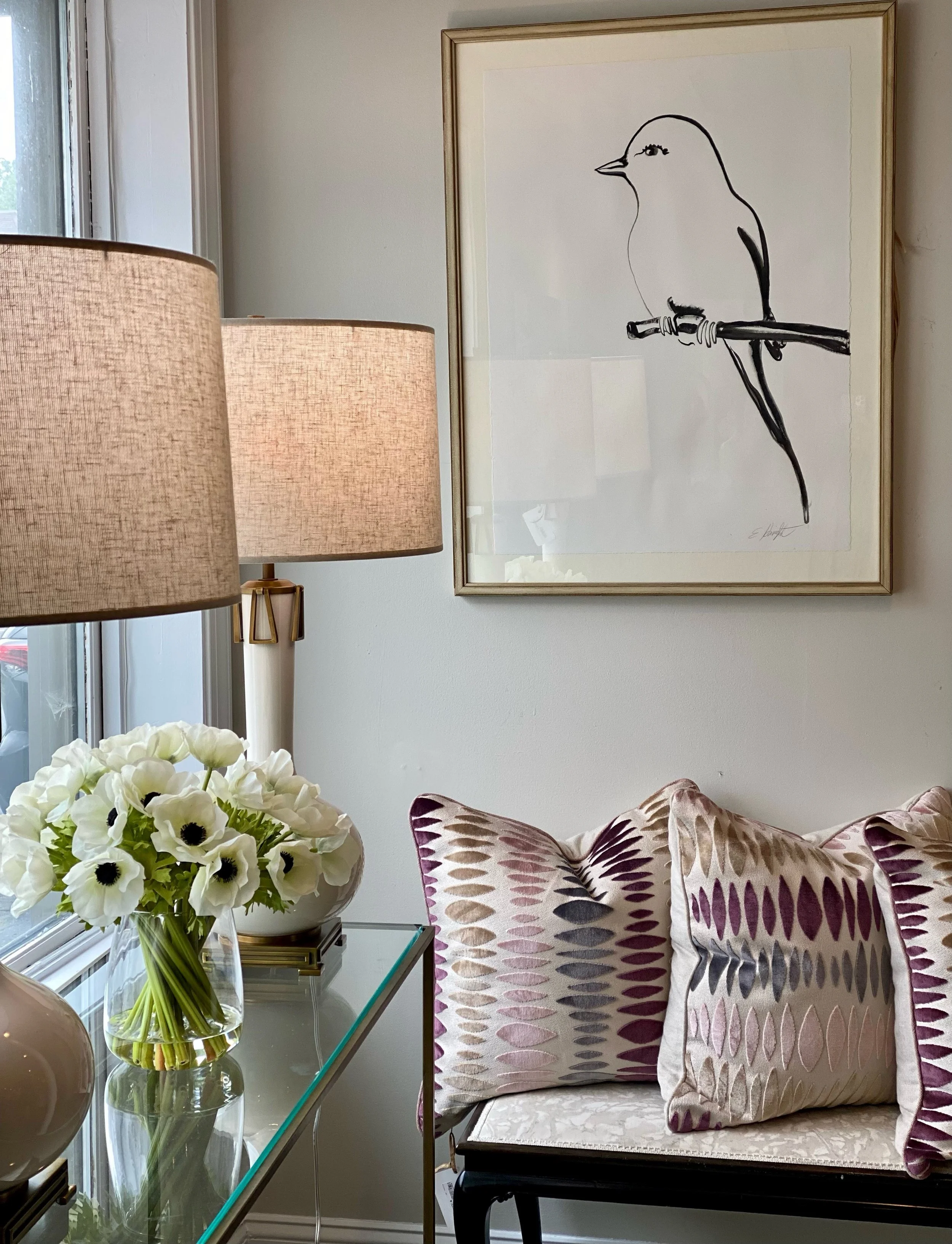 Interior of a room with a table holding a large glass vase with white flowers, a succulent, and brown vases. There is a window with white trim, a framed line drawing of a bird on the wall, and a patterned pillow and cushion on a chair.