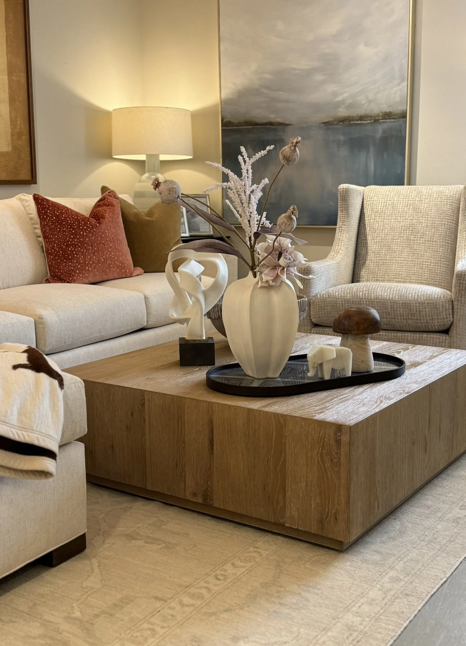 Living room with a beige sofa, a beige armchair, a wooden coffee table with decorative items, a large abstract painting, a lamp on the side table, and a floral arrangement in a white vase.