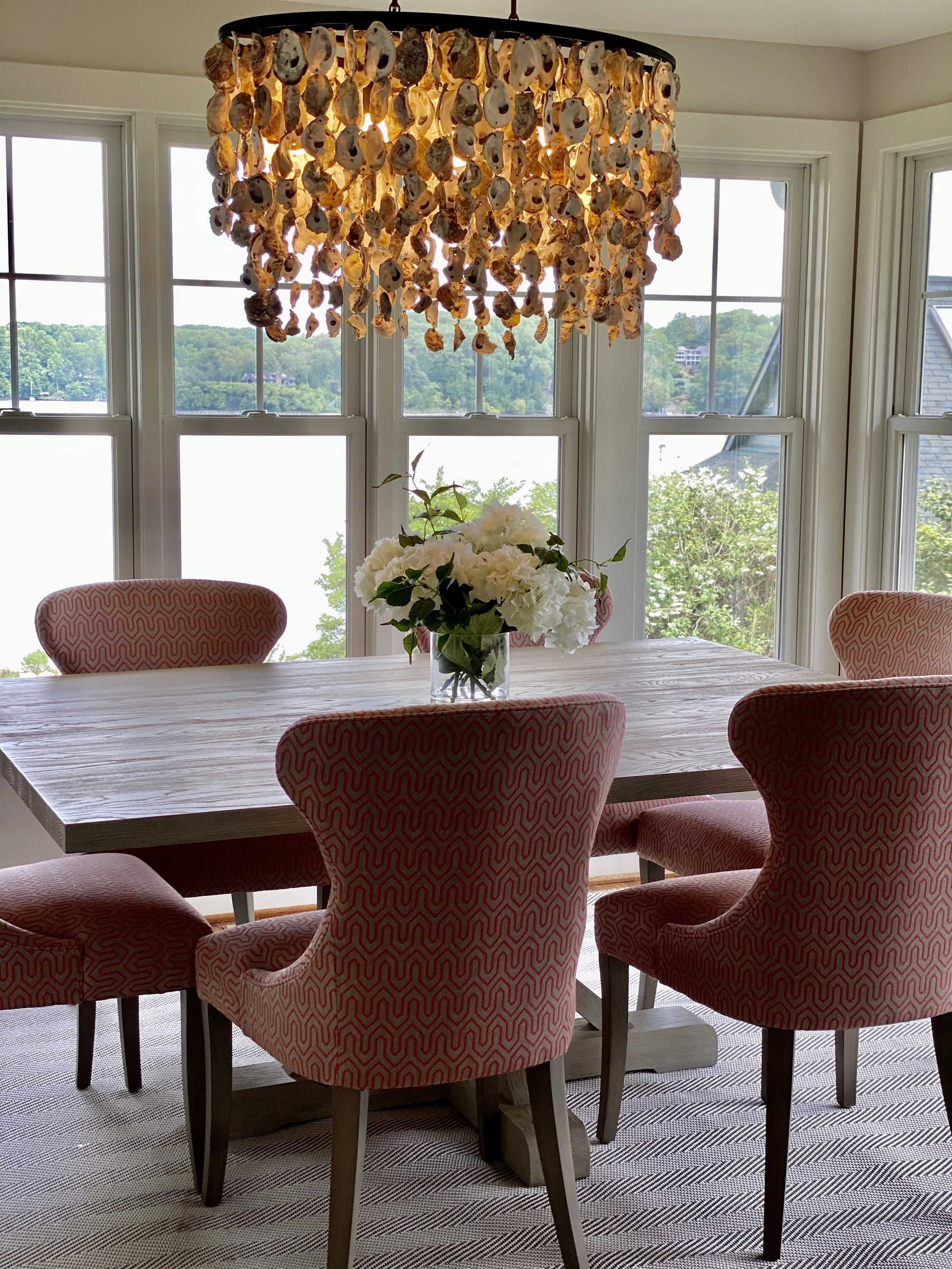 A dining room with a wooden table surrounded by six pink and white patterned chairs. A vase with white flowers sits on the table. The room has large windows with views of green trees and a river outside, and a chandelier made of seashells hanging above the table.