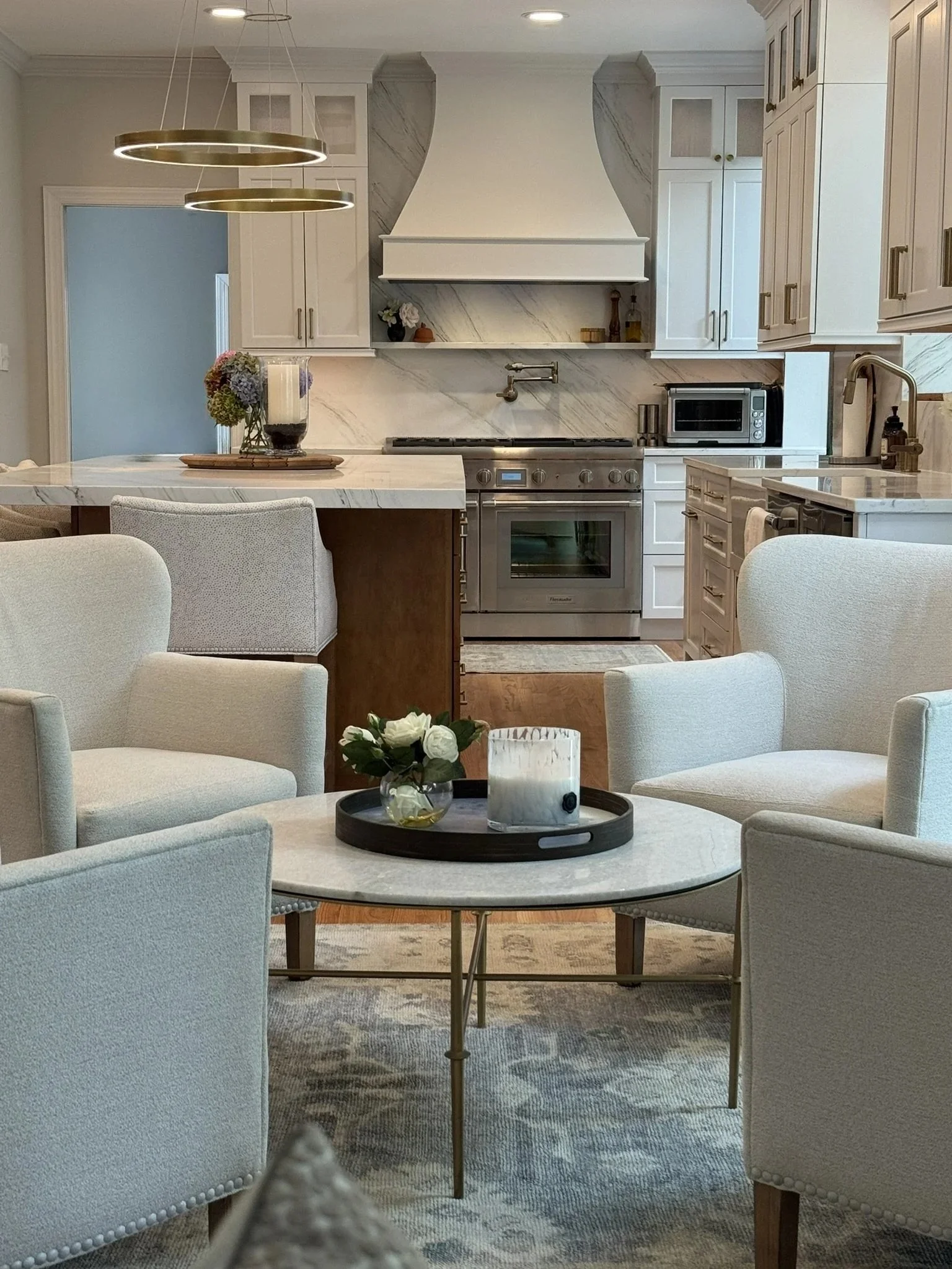 A modern kitchen area with white cabinets, marble backsplash, stainless steel oven, microwave, and a round table with beige upholstered chairs. There are decorative items like flowers and candles on the table and counter. Two gold pendant lights hang above the dining table.
