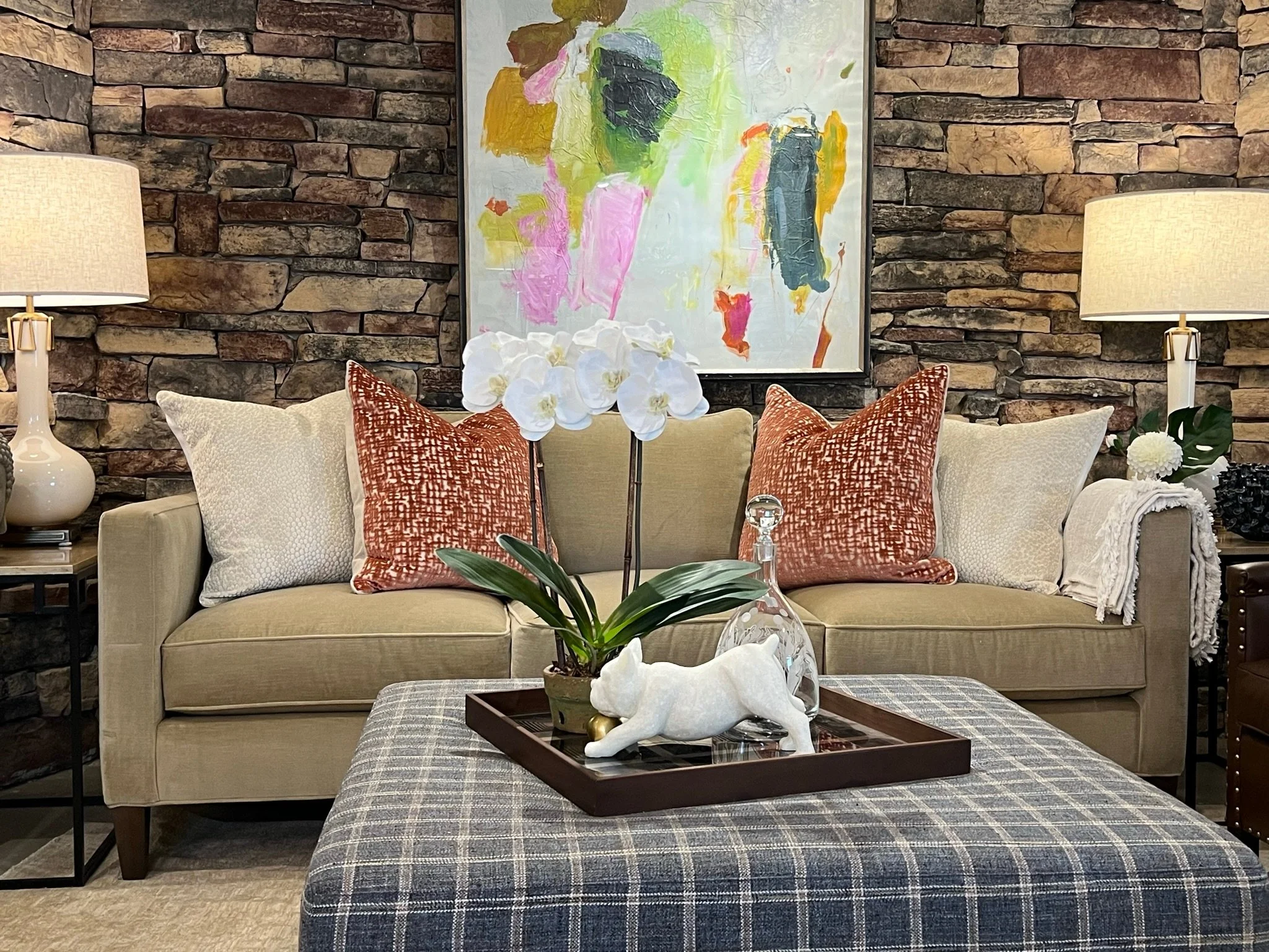 Living room with stone wall, colorful abstract painting, beige sofa with white and rust pillows, table with white orchid plant in a decorative pot, lamps on side tables.