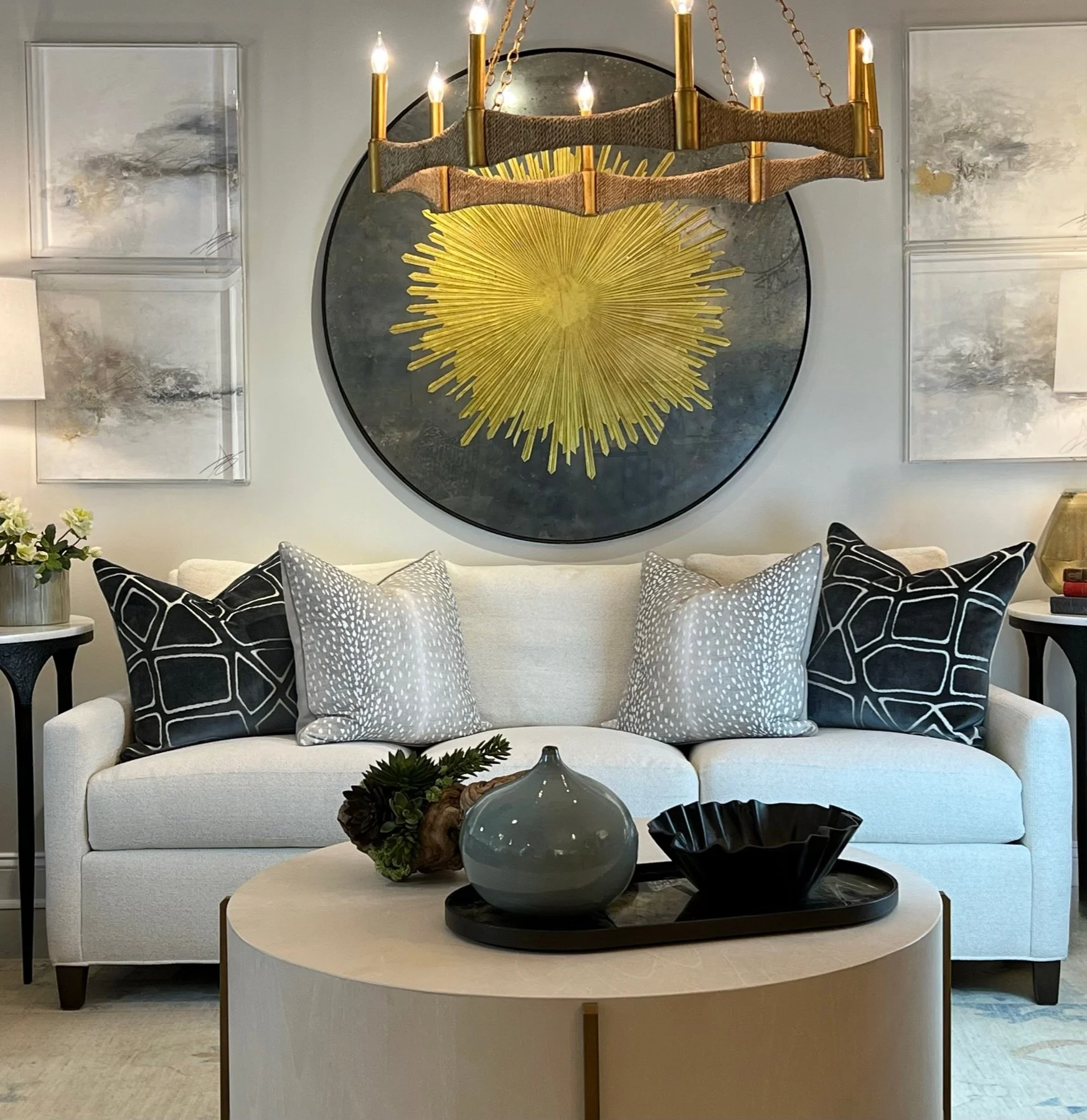 The image depicts a modern living room with a white sofa adorned with patterned black and white cushions. In front of the sofa is a oval, light-colored coffee table with decorative objects including a green plant, a gray vase, and a black bowl. Behin
