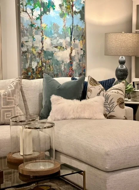 A living room with a sectional sofa decorated with various pillows. Behind the sofa, there is a colorful abstract painting and a table lamp with a textured shade.