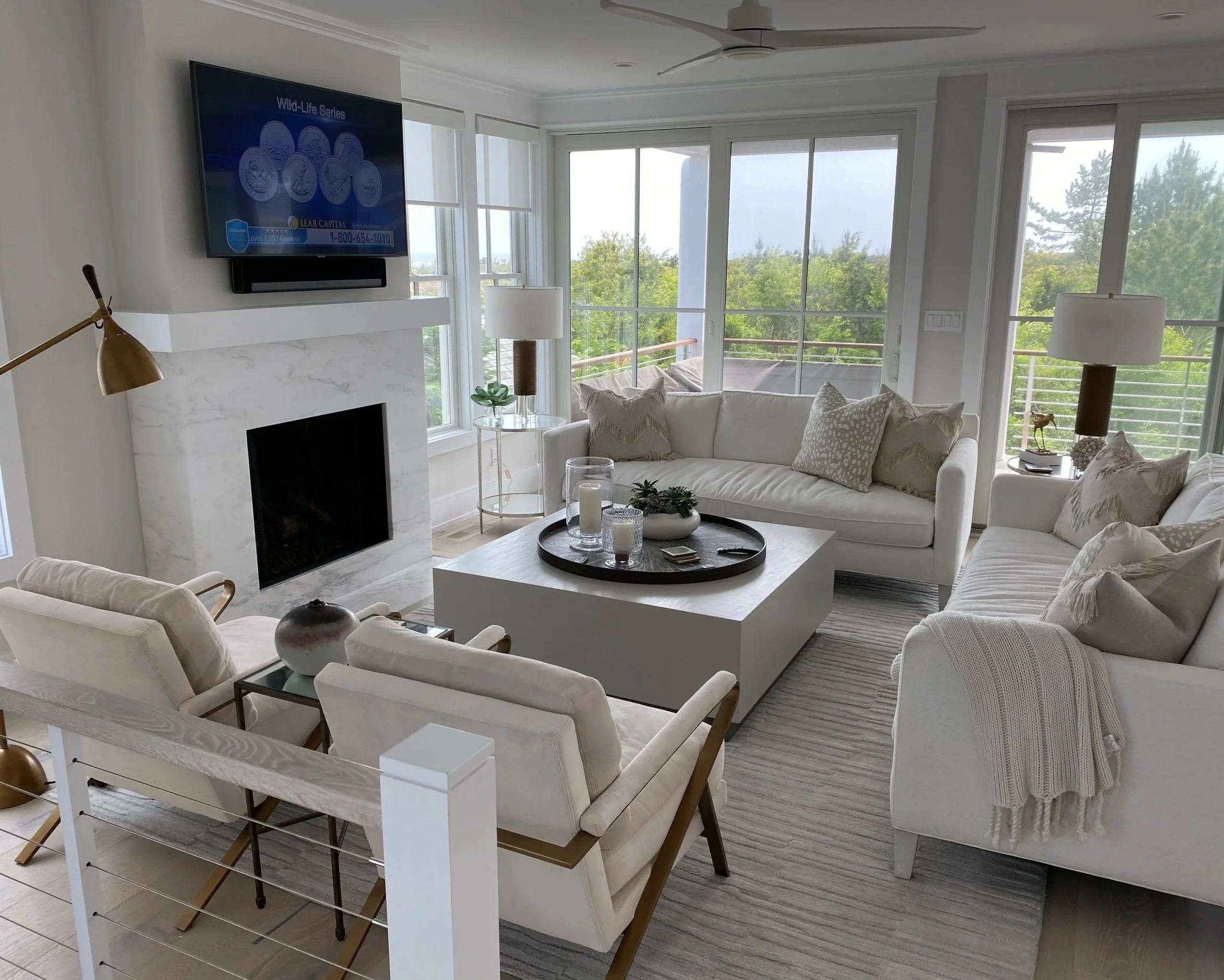 Bright living room with large windows and white walls, featuring a white sofa with decorative pillows, a marble fireplace, a round coffee table with candles and decor, a side table with plants, lamps, and a wall-mounted flat-screen TV.