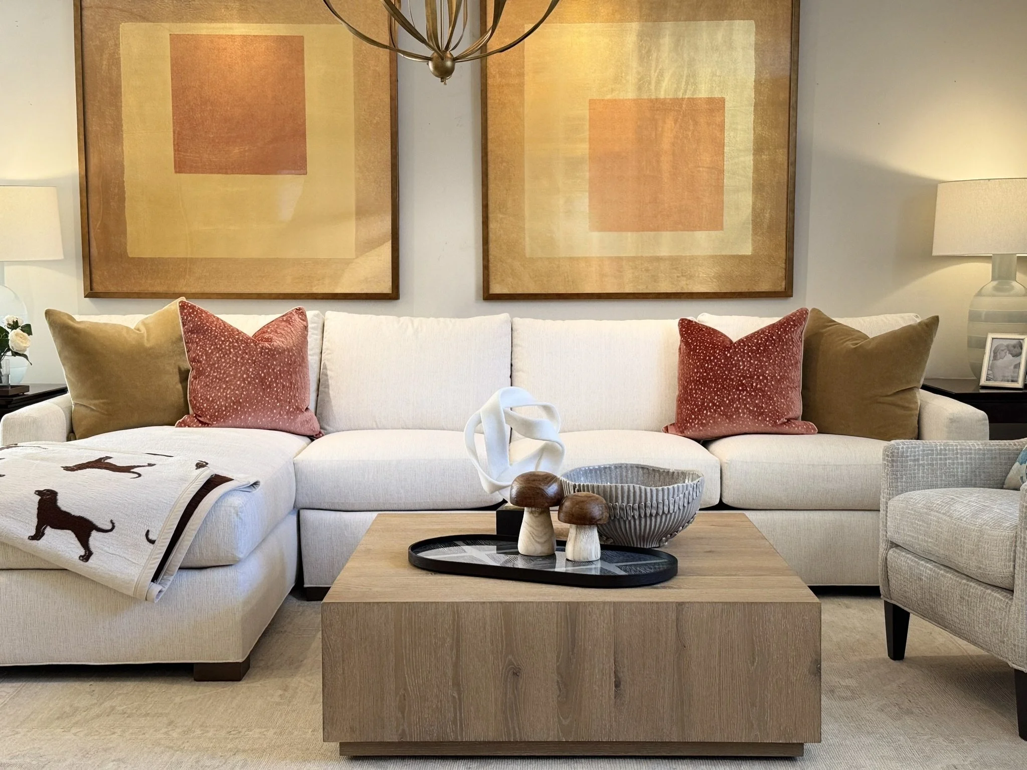 Living room with a white sofa decorated with pink and tan pillows, a wooden coffee table with decorative items, and large abstract art paintings on the wall behind the sofa.