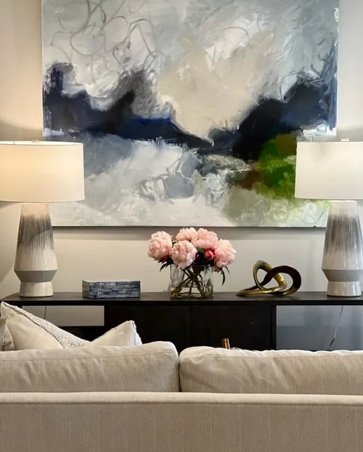 Living room with two table lamps, a large abstract painting, a vase of pink flowers, and decorative objects on a black console table, with a beige sofa in the foreground.
