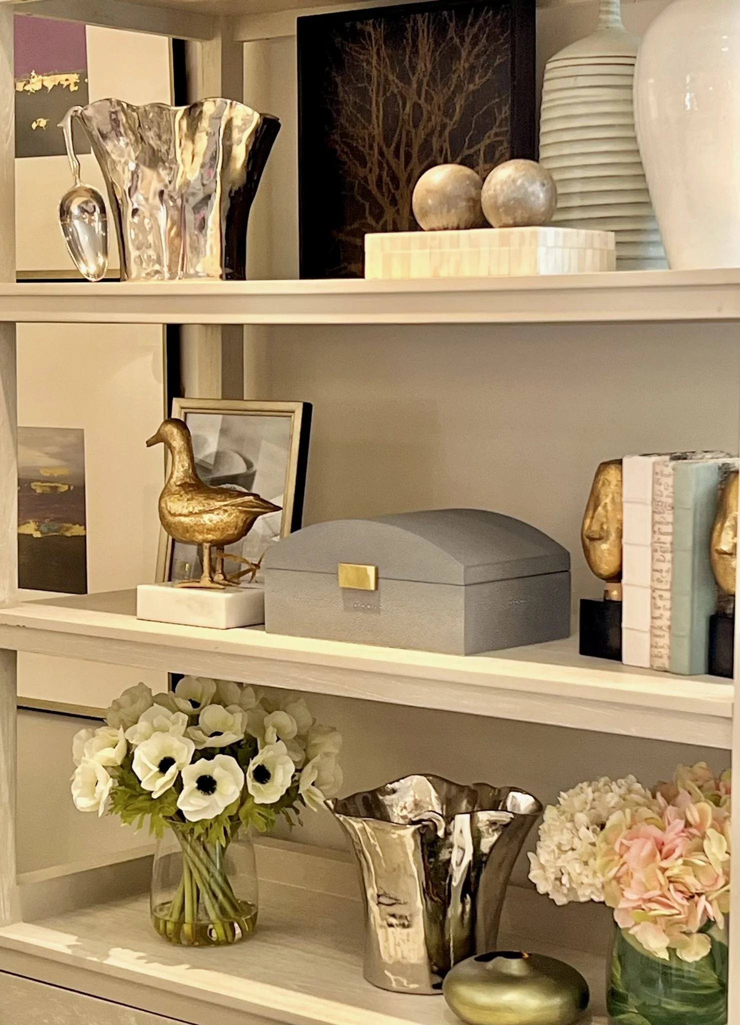 A white shelf decorated with vases, artwork, and flowers. The top shelf holds a shiny metallic vase, black box, framed artwork of a tree, and several white and beige vases. The bottom shelf features a glass vase with white flowers, a black and white 