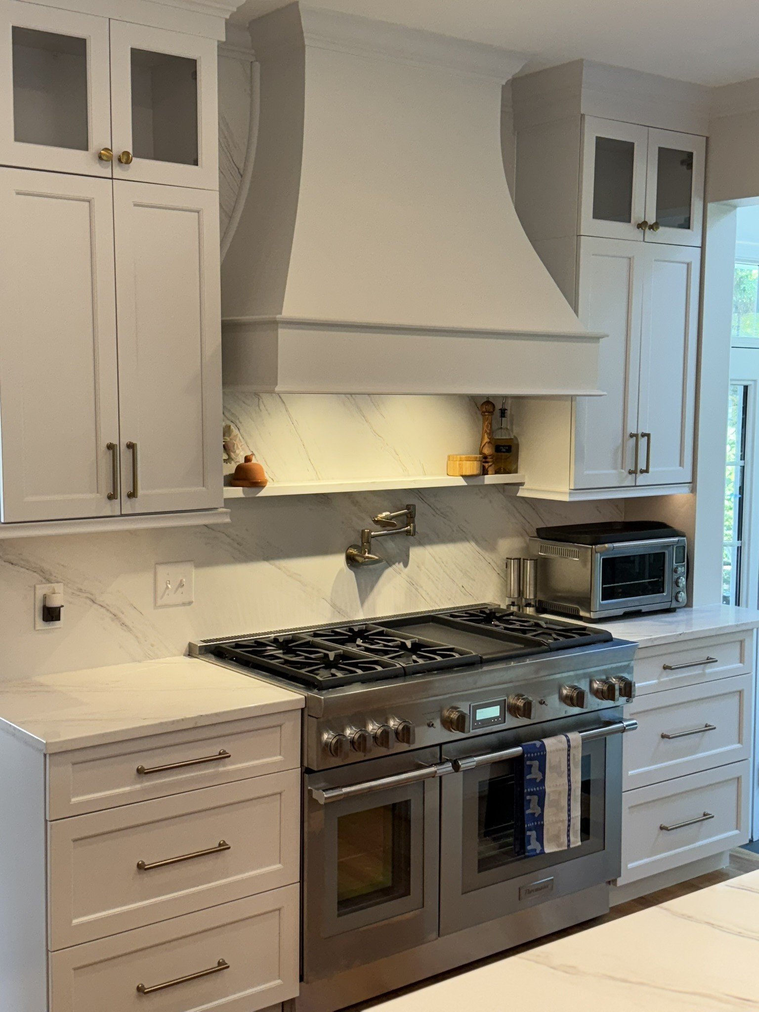 White kitchen with built-in cabinets, stainless steel oven and stove, marble backsplash, and some small kitchen items on the counter.