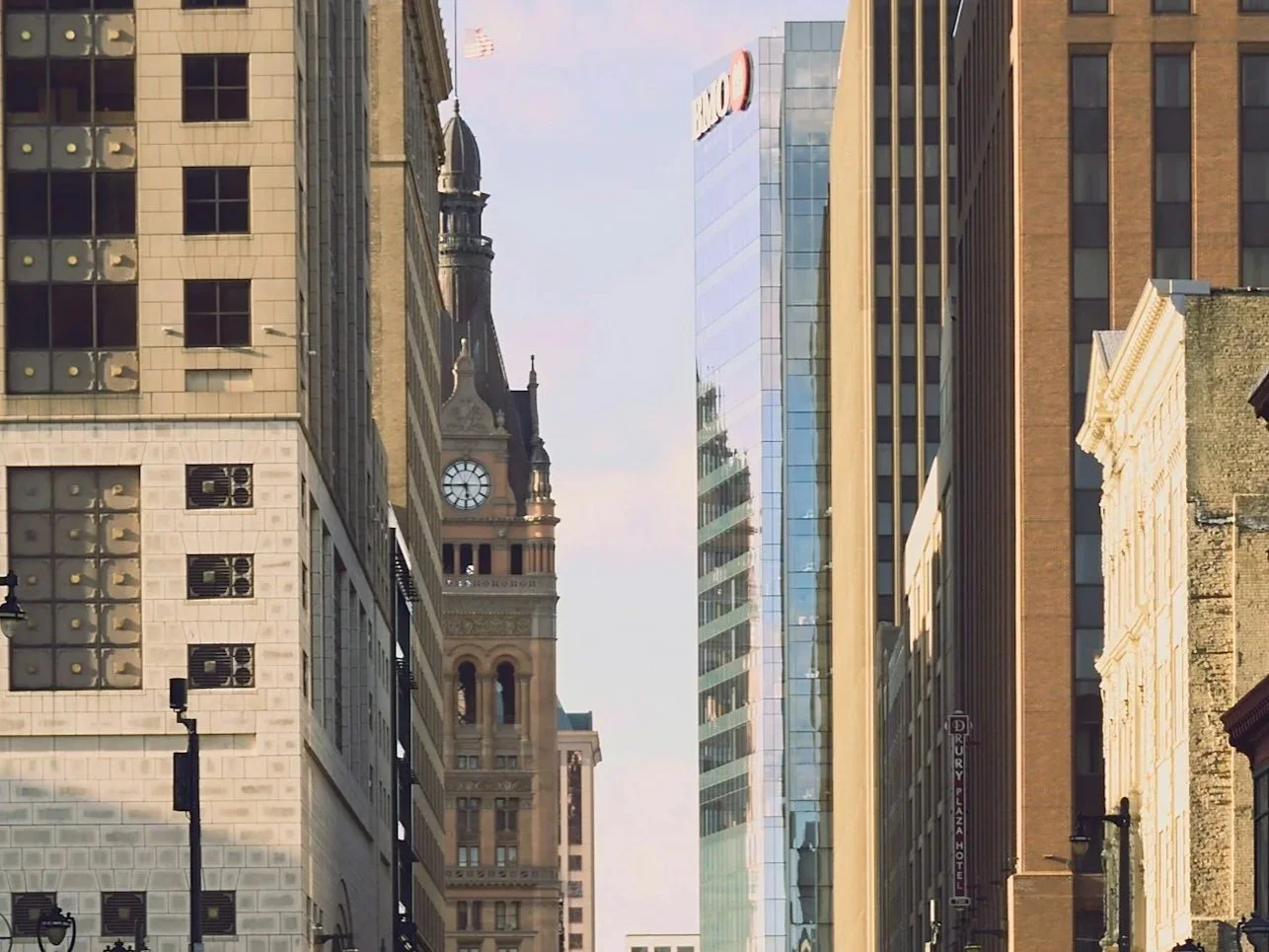 City street with tall historic and modern buildings, including a clock tower and glass skyscraper, under a cloudy sky.