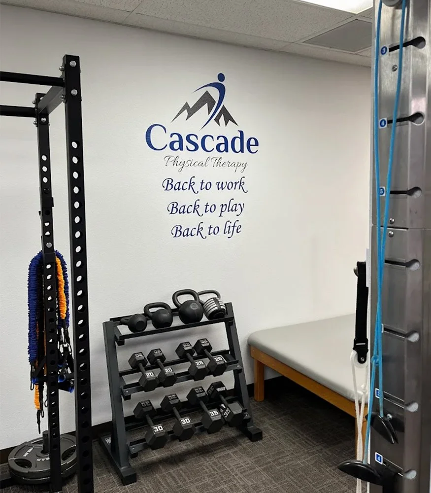 Interior view of a physical therapy room with workout equipment, including dumbbells, kettlebells, resistance bands, and a therapy table, with a motivational phrase on the wall.