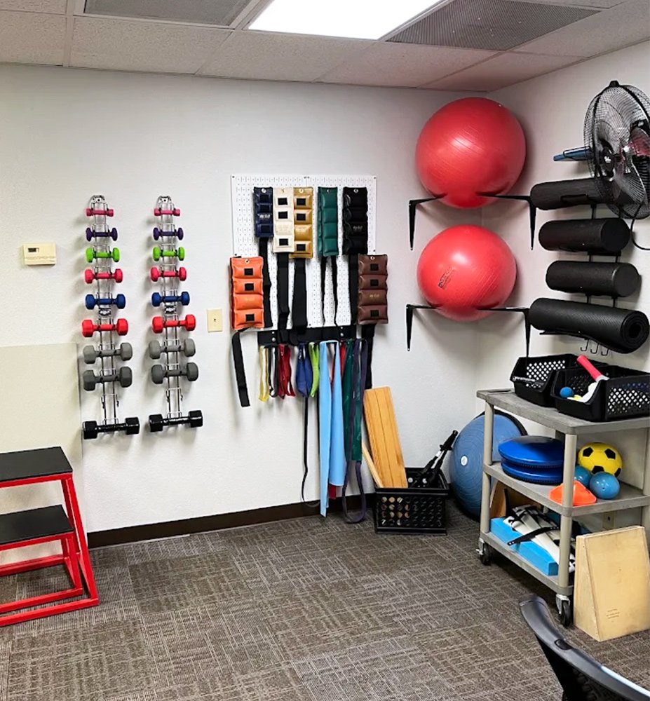 Workout room with colorful dumbbells on wall, resistance bands hanging on a pegboard, large red exercise balls on wall mounts, foam rollers, a fan, and various exercise equipment on a cart, against a beige carpet and ceiling tiles.