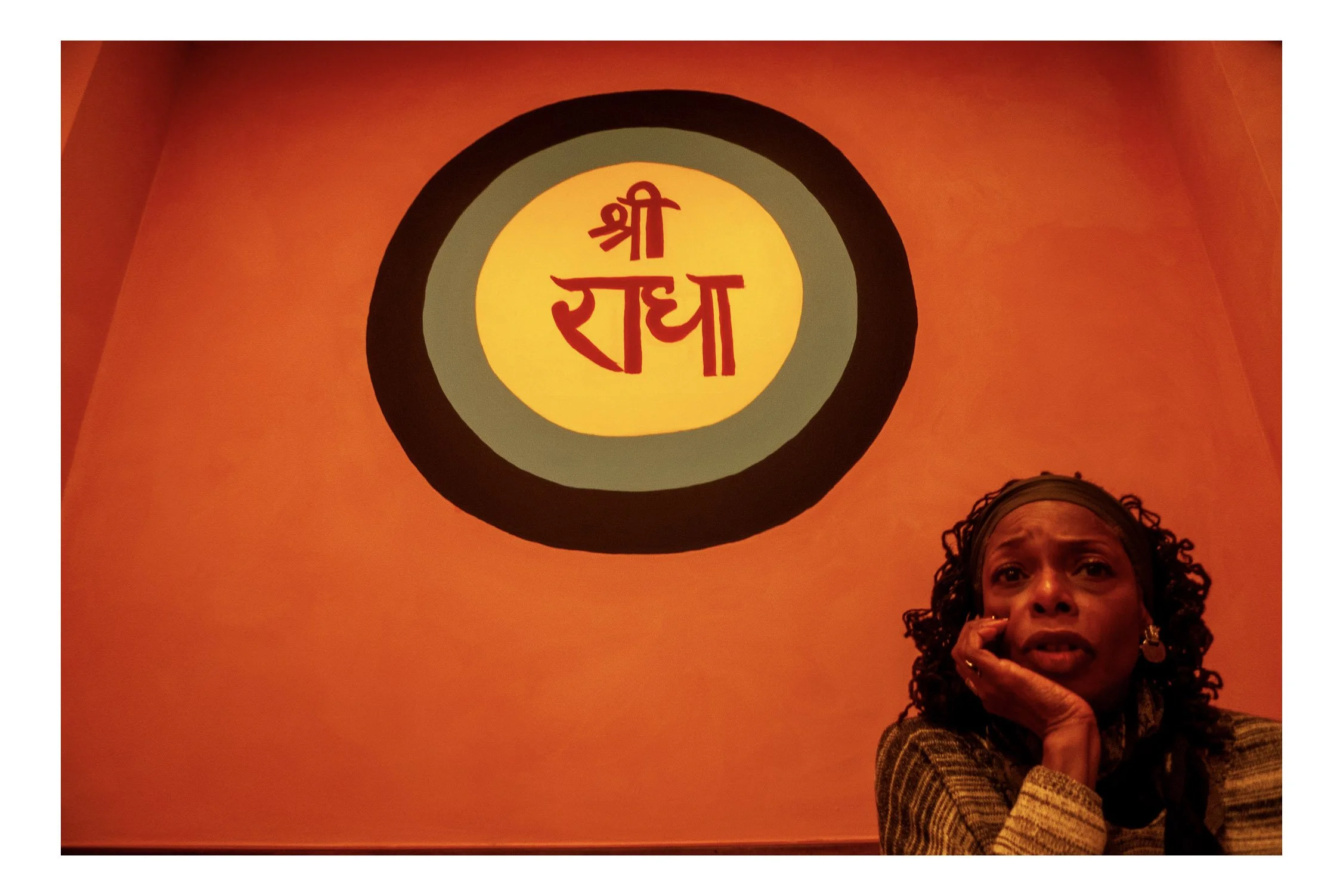 A woman is sitting indoors with an orange background and a circular decorative piece with red Hindi text that reads 'Shraddha' in the center. The woman is wearing a checkered blazer, has curly hair, and is holding her hand near her face.