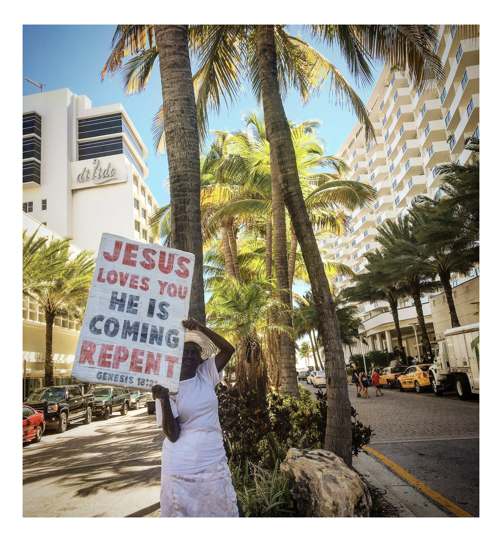 Person holding a sign that reads 'JESUS LOVES YOU HE IS COMING' standing among palm trees on a city street with high-rise buildings and parked cars.