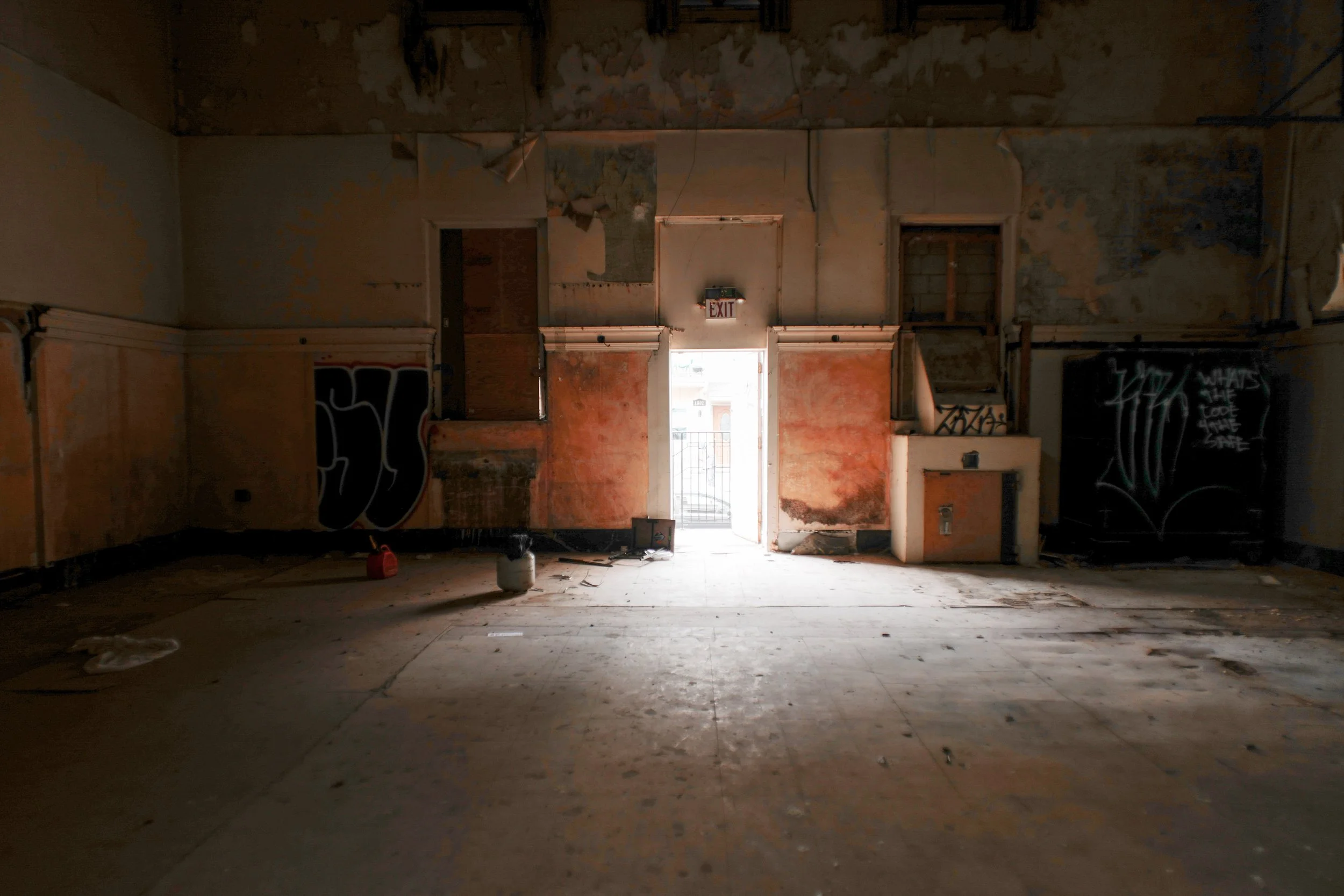 An abandoned and derelict room with peeling paint, graffiti on the walls, and broken or missing ceiling panels. Light enters through a door at the far end, which leads outside, creating a silhouette effect.