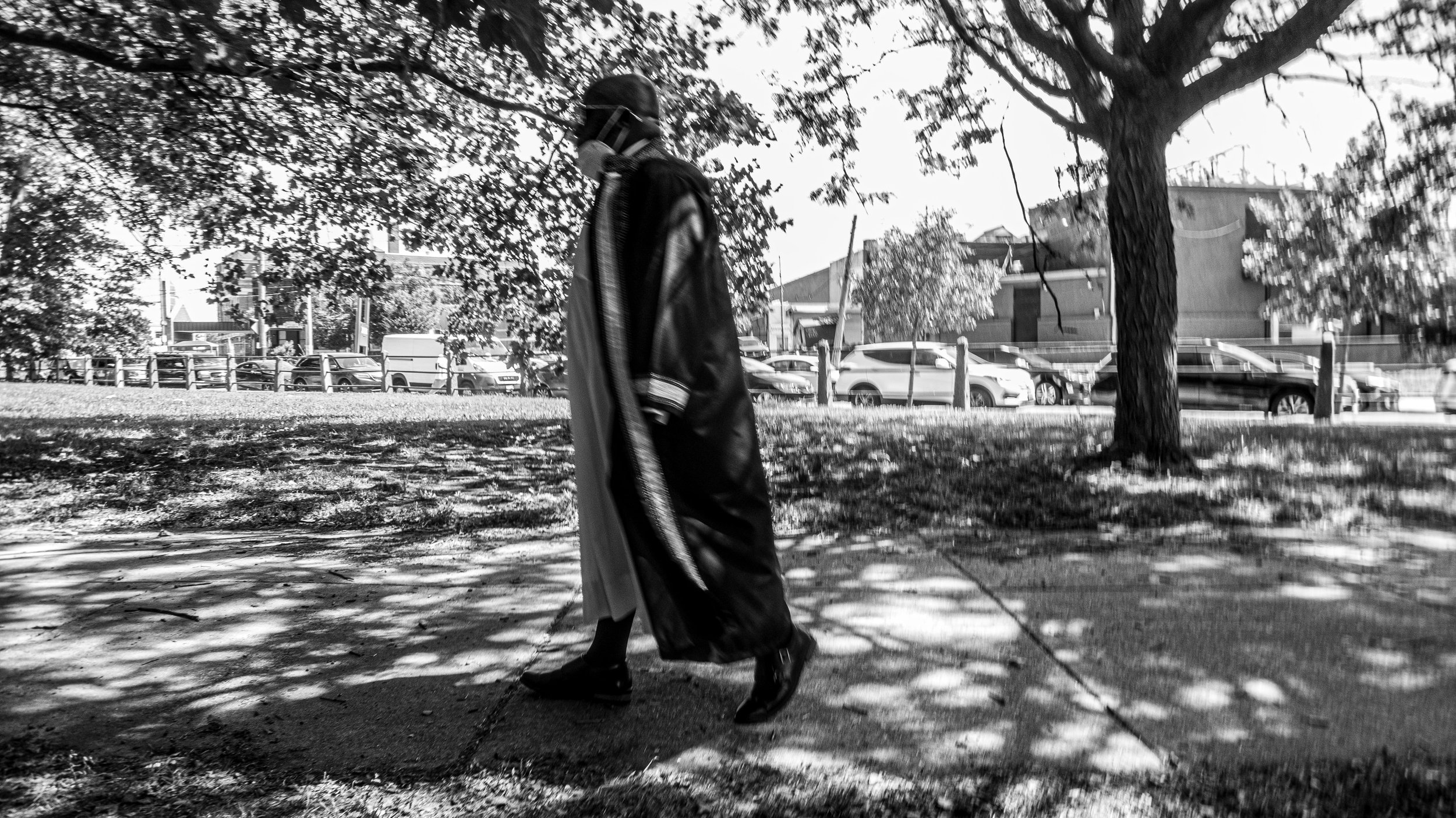 A person in a black coat, boots, and face mask walking in a park with trees and parked cars in the background.